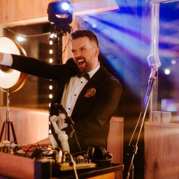 Kultowa Firma Marcin Jesionek - Uśmiechnięty DJ w smokingu, z muszką, miksuje muzykę na konsolecie, oświetlony reflektorami i dekoracyjnymi lampkami podczas imprezy.