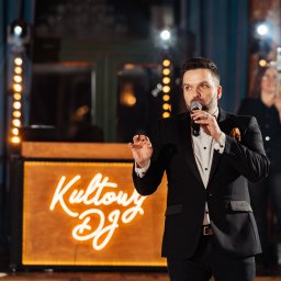 Kultowa Firma Marcin Jesionek - Mężczyzna w garniturze przemawia do mikrofonu na tle podświetlonego napisu 'Kultowy DJ', gitara i oświetlenie sceniczne w tle, osoba filmująca smartfonem.