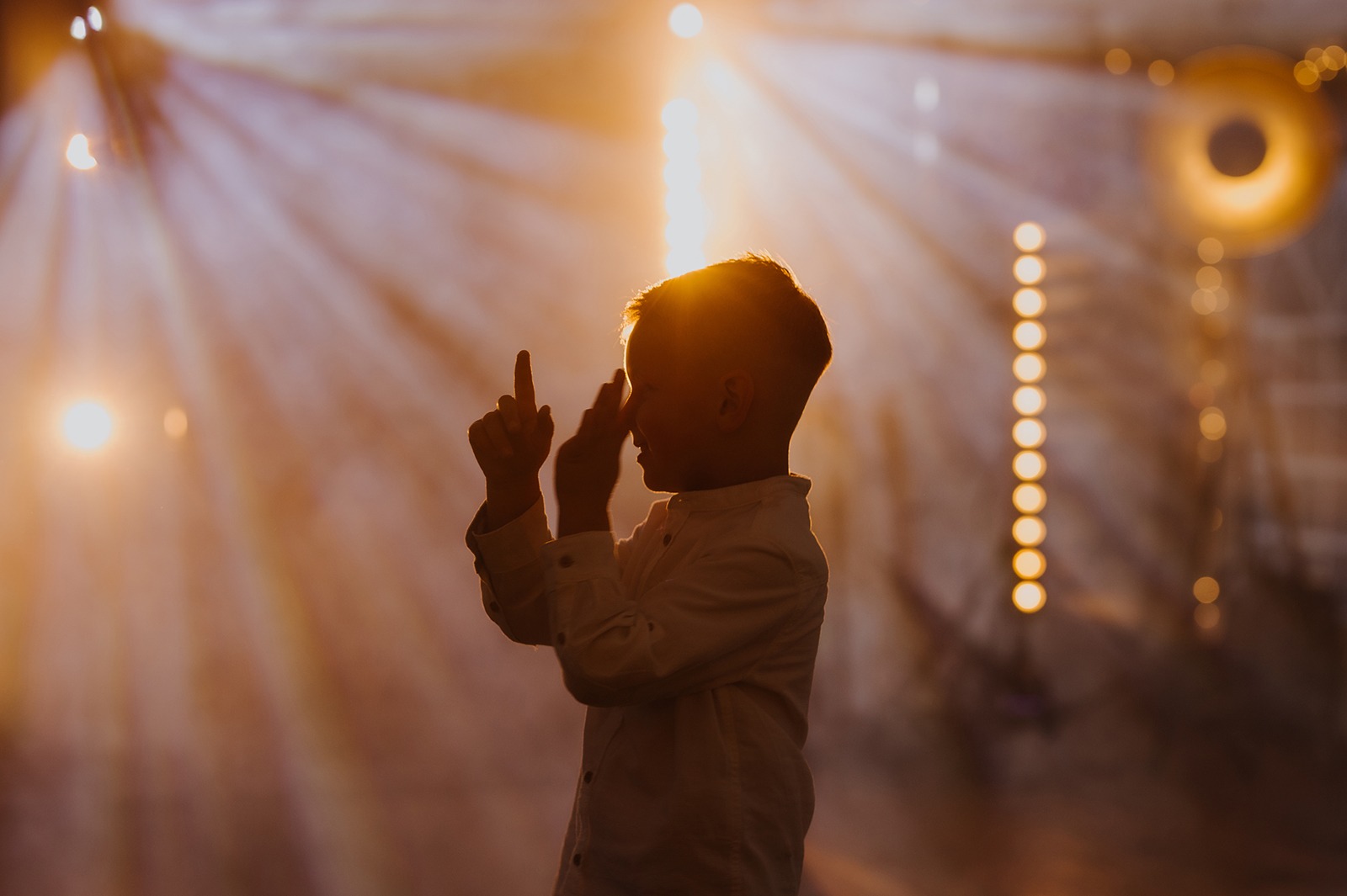 Silueta chłopca w białej koszuli na tle sceny z reflektorami. Dziecko wskazuje palcami w górę, oświetlone ciepłym światłem. Koncertowe oświetlenie w tle.