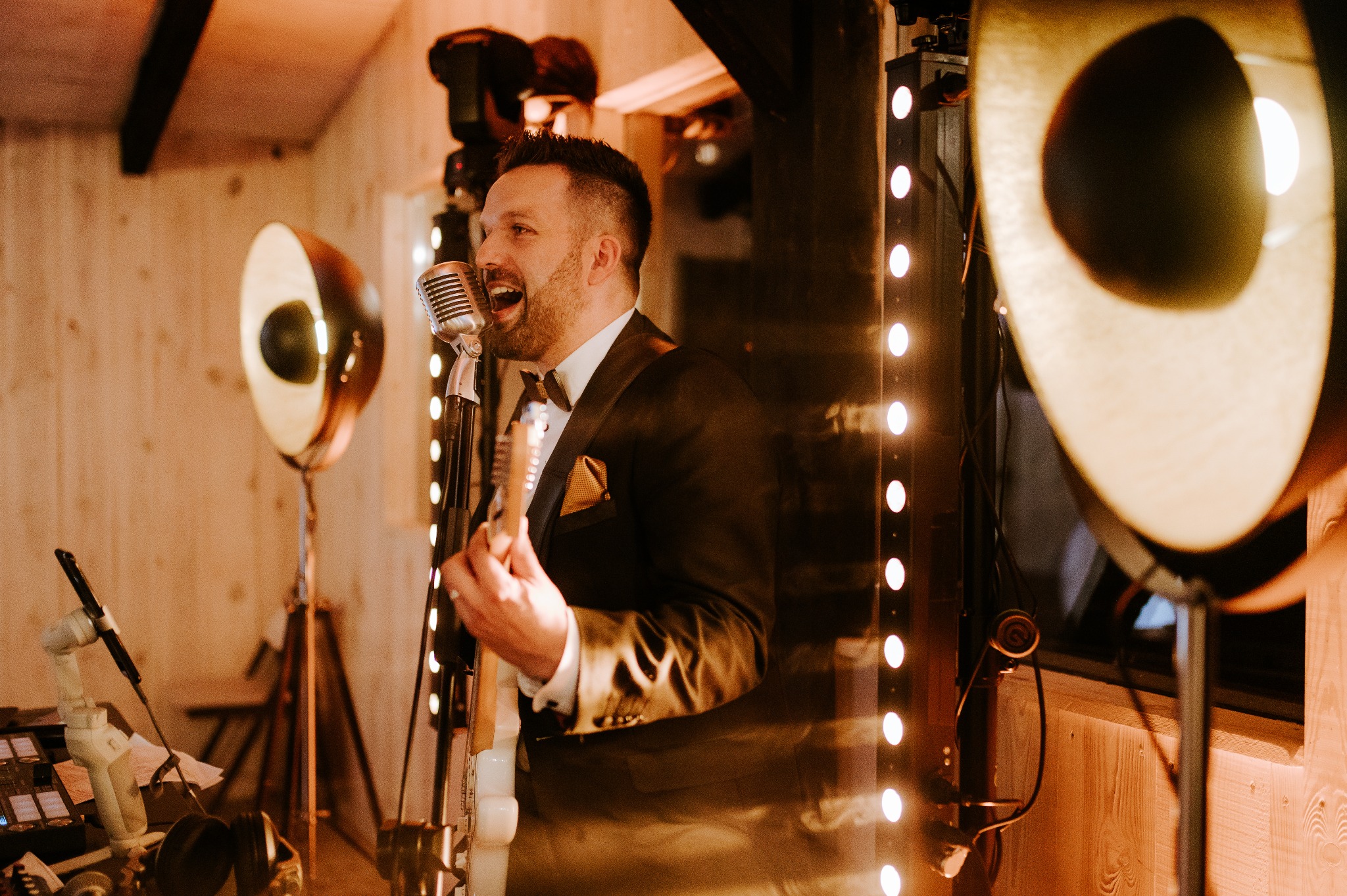 Uśmiechnięty muzyk w smokingu gra na gitarze i śpiewa do mikrofonu na tle oświetlenia scenicznego w ciepłej tonacji.