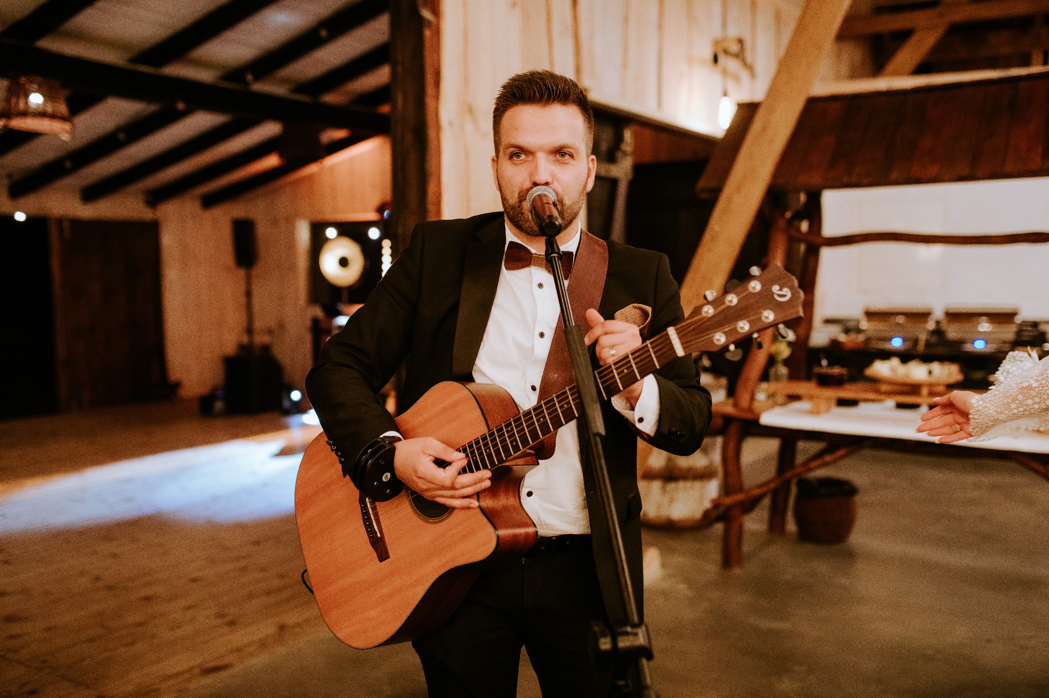 Elegancki muzyk w smokingu gra na gitarze akustycznej i śpiewa do mikrofonu na tle rustykalnej sali z drewnianymi elementami dekoracyjnymi i bufetem.