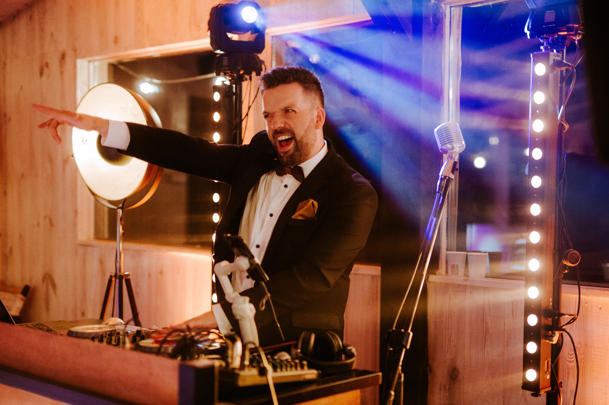 Uśmiechnięty DJ w smokingu, z muszką, miksuje muzykę na konsolecie, oświetlony reflektorami i dekoracyjnymi lampkami podczas imprezy.