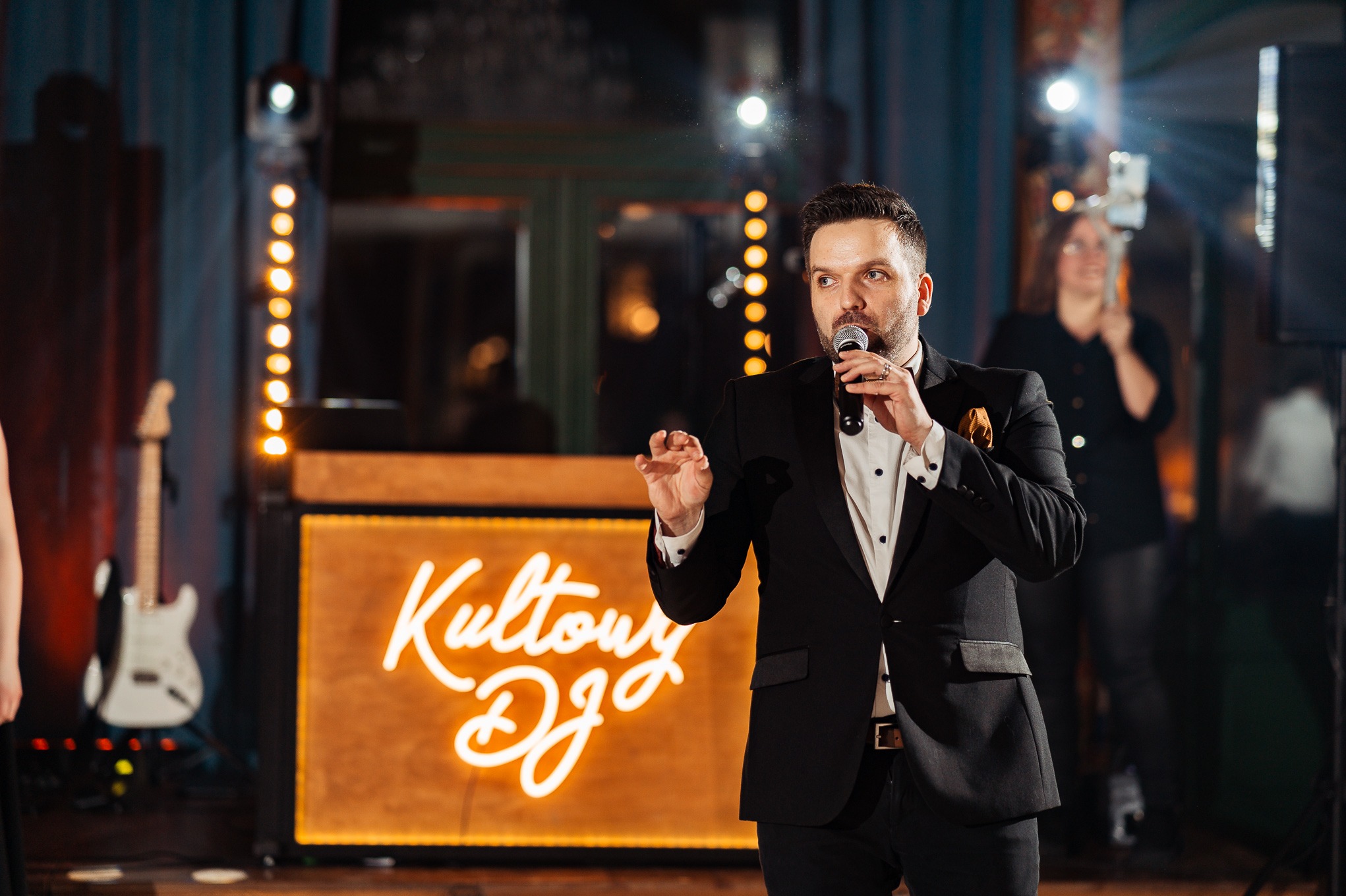 Mężczyzna w garniturze przemawia do mikrofonu na tle podświetlonego napisu 'Kultowy DJ', gitara i oświetlenie sceniczne w tle, osoba filmująca smartfonem.