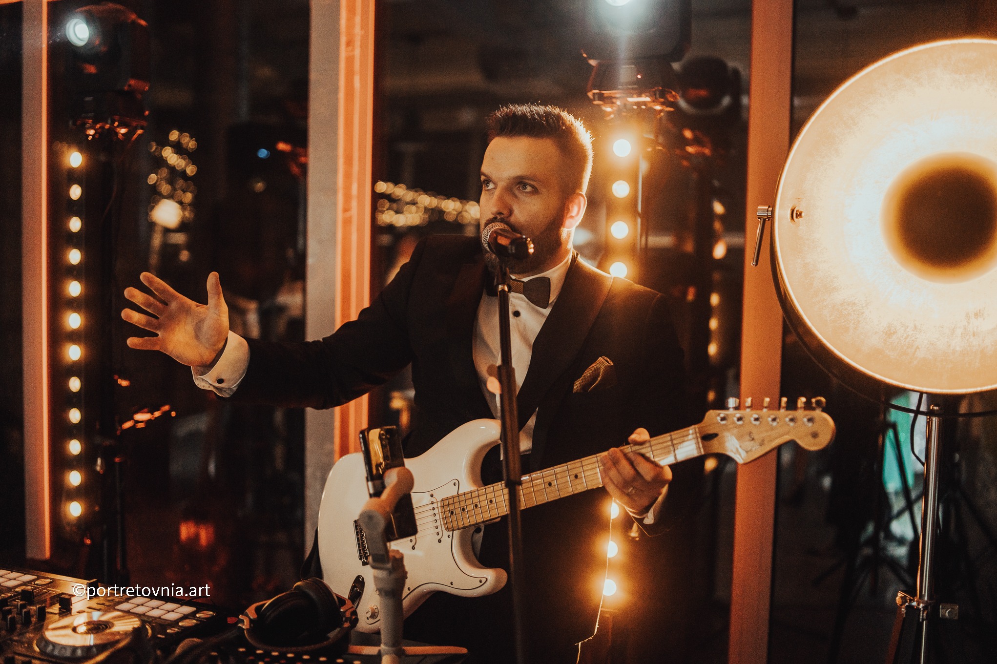 Elegancki muzyk w smokingu gra na białej gitarze elektrycznej i śpiewa do mikrofonu na tle dekoracji świetlnych. Widoczny sprzęt DJ-ski.