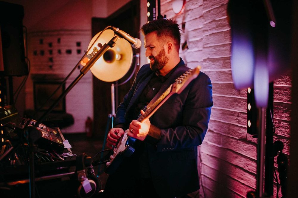 Muzyk z brodą w ciemnym garniturze gra na gitarze elektrycznej na tle ceglanej ściany oświetlonej czerwonym światłem. Widoczny mikrofon i sprzęt muzyczny.