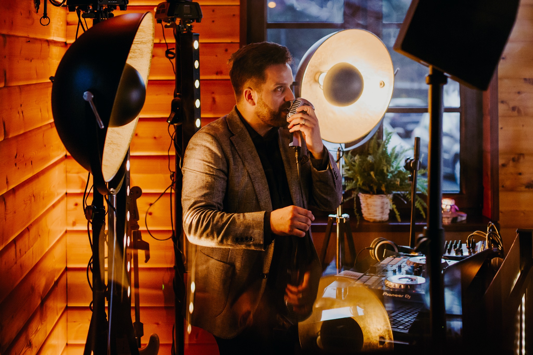 Wokalista w tweedowej marynarce śpiewa do mikrofonu retro na tle drewnianej ściany, oświetlenia scenicznego i sprzętu DJ-skiego Pioneer DJ.