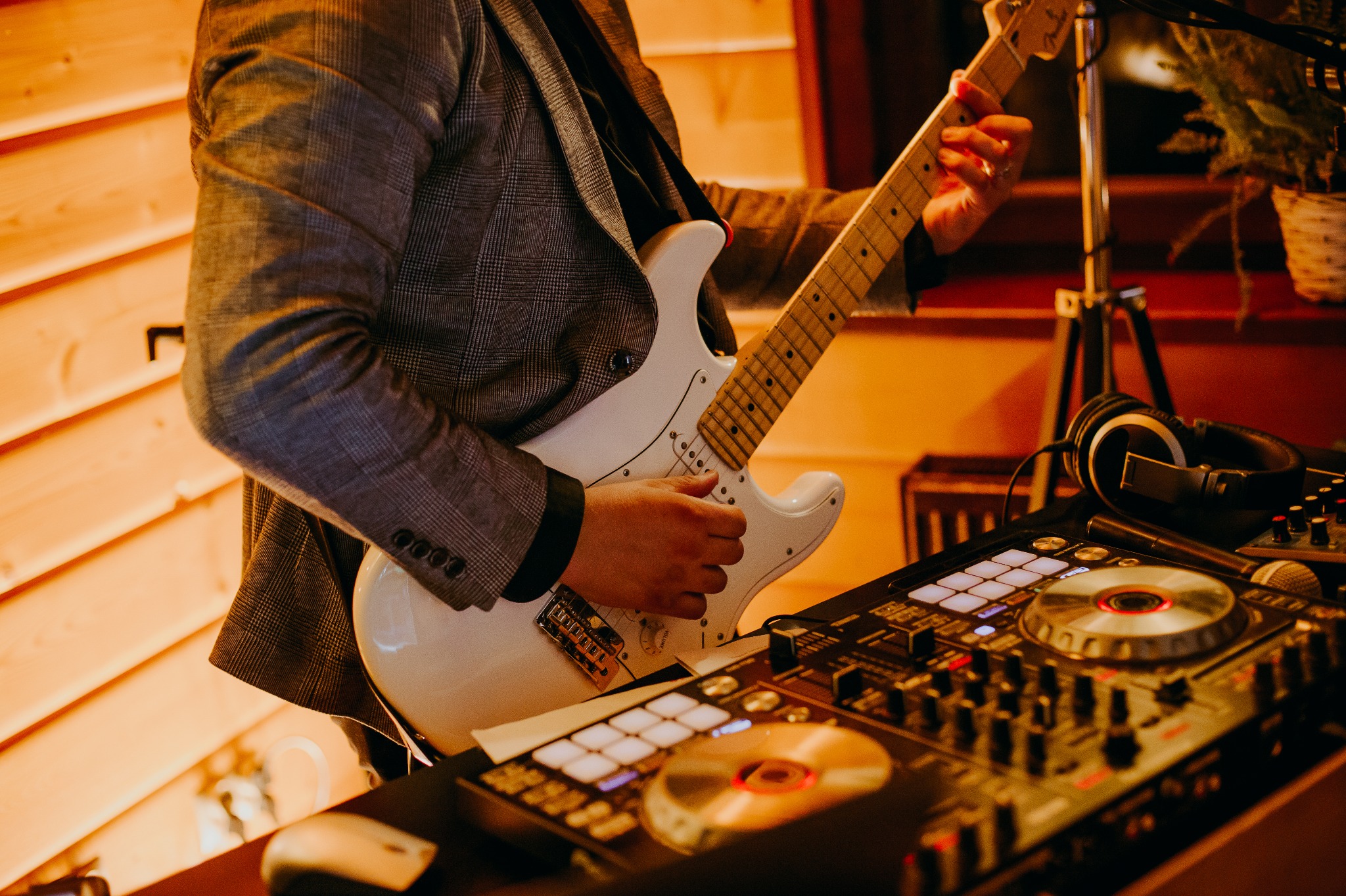 Gitarzysta w tweedowej marynarce gra na białej gitarze elektrycznej, obok sprzęt DJ-ski. Ciepłe oświetlenie sceniczne, drewniane panele w tle.