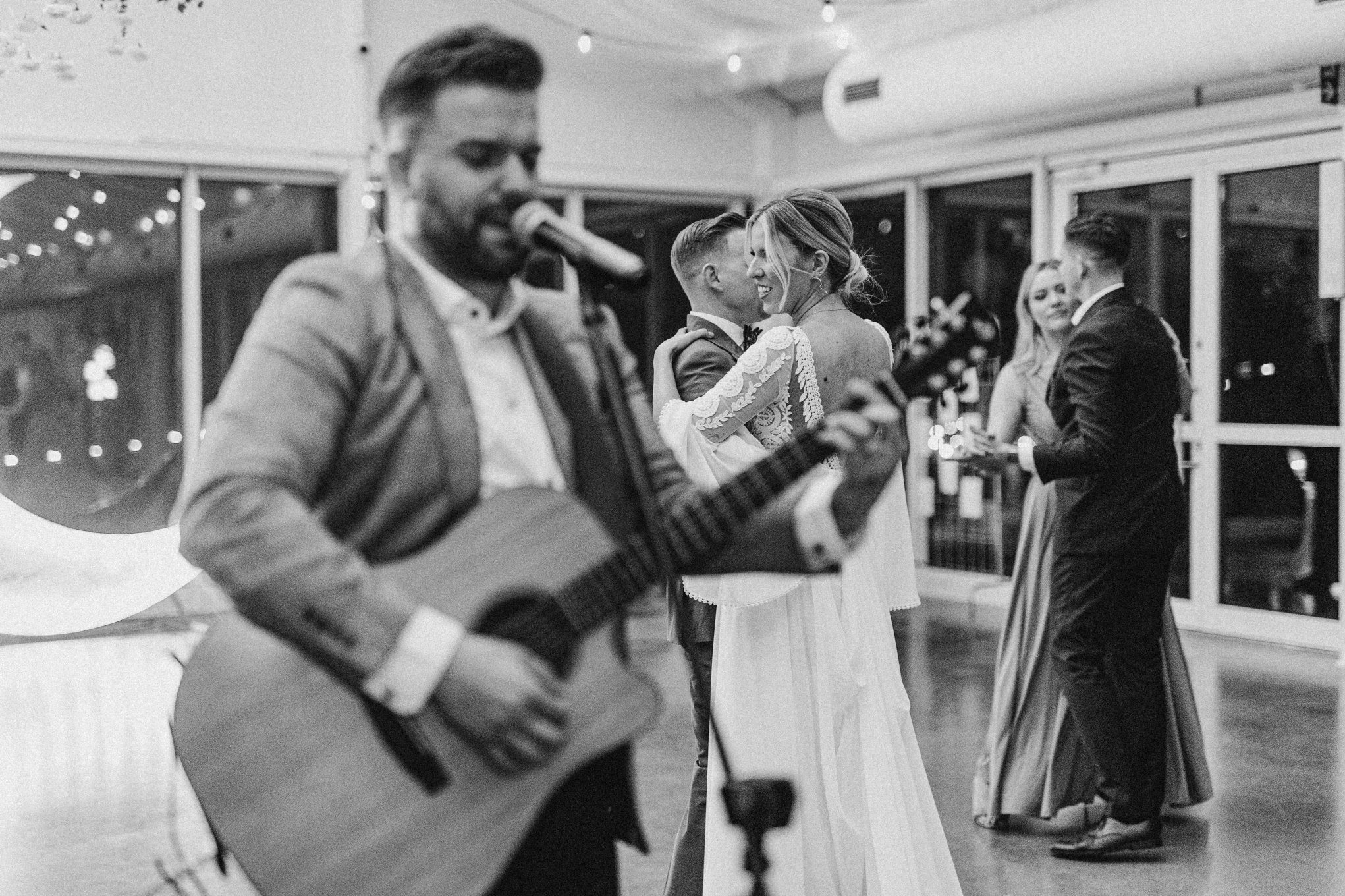 Czarno-białe zdjęcie muzyka grającego na gitarze i śpiewającego na weselu, para młoda tańczy w tle, a goście weselni obserwują.