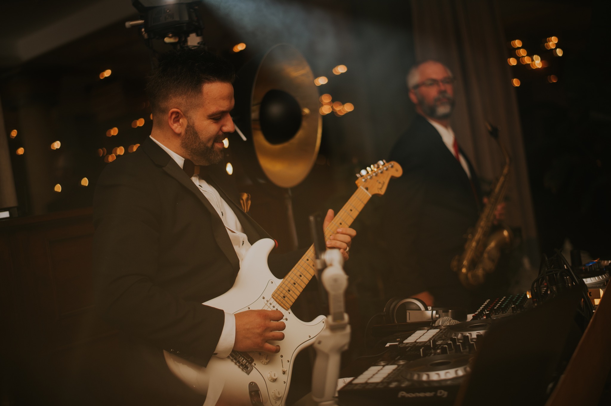 Elegancki gitarzysta w akcji, z saksofonistą w tle i sprzętem DJ-skim na pierwszym planie, tworzący unikalną atmosferę muzyczną na evencie.