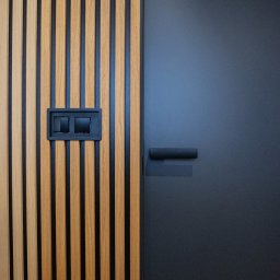 Mariusz Guzera - Panel ścienny z drewnianych listew w odcieniach brązu, z czarnymi wstawkami i gniazdkiem elektrycznym, sąsiadujący z gładkimi, grafitowymi drzwiami z minimalistyczną klamką.