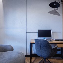 Mariusz Guzera - Nowoczesne wnętrze biurowe z teksturowaną ścianą, drewnianym biurkiem z czarną ramą, szarym fotelem i lampą. Minimalistyczny design z akcentami drewna.