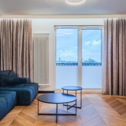 Mariusz Guzera - Salon z parkietem w jodełkę, niebieską sofą i okrągłymi stolikami. Widok na miasto przez okno z plisowanymi roletami. Nowoczesne oświetlenie i minimalistyczny wystrój.