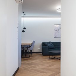 Mariusz Guzera - Jasne parkietowe panele ułożone w jodełkę, widok z korytarza na salon z sofą i jadalnię z czarnymi lampami. Minimalistyczne wnętrze w odcieniach bieli i szarości.