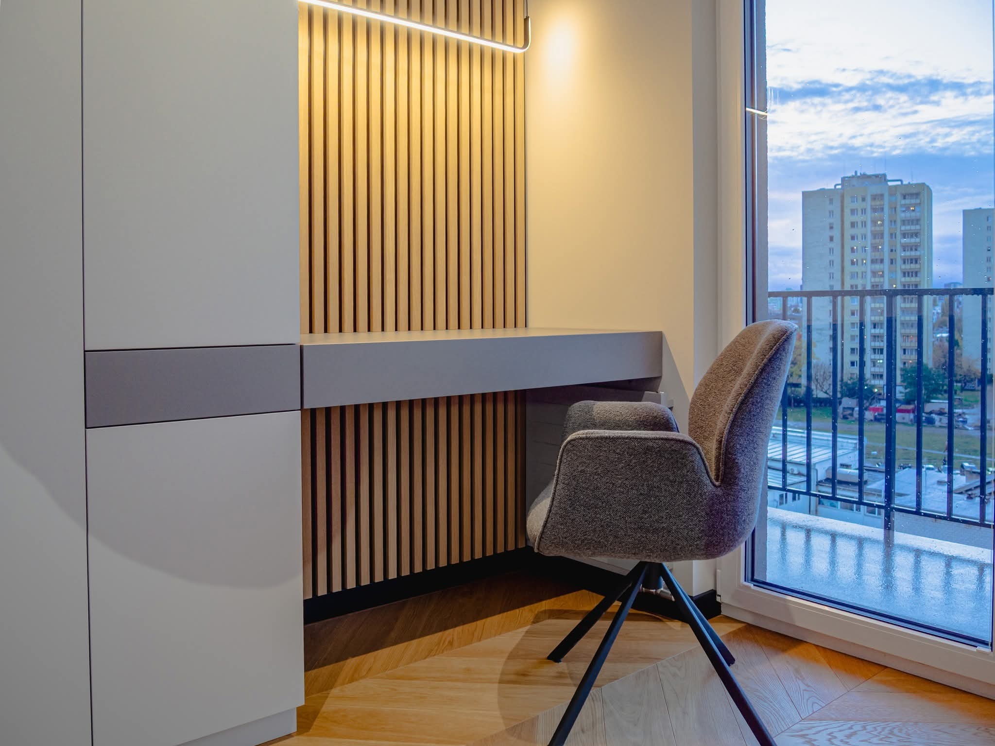 Nowoczesny pokój z biurkiem, drewnianą ścianką działową i krzesłem na tle okna z widokiem na miasto. Parkiet ułożony w jodełkę. Minimalistyczny design.