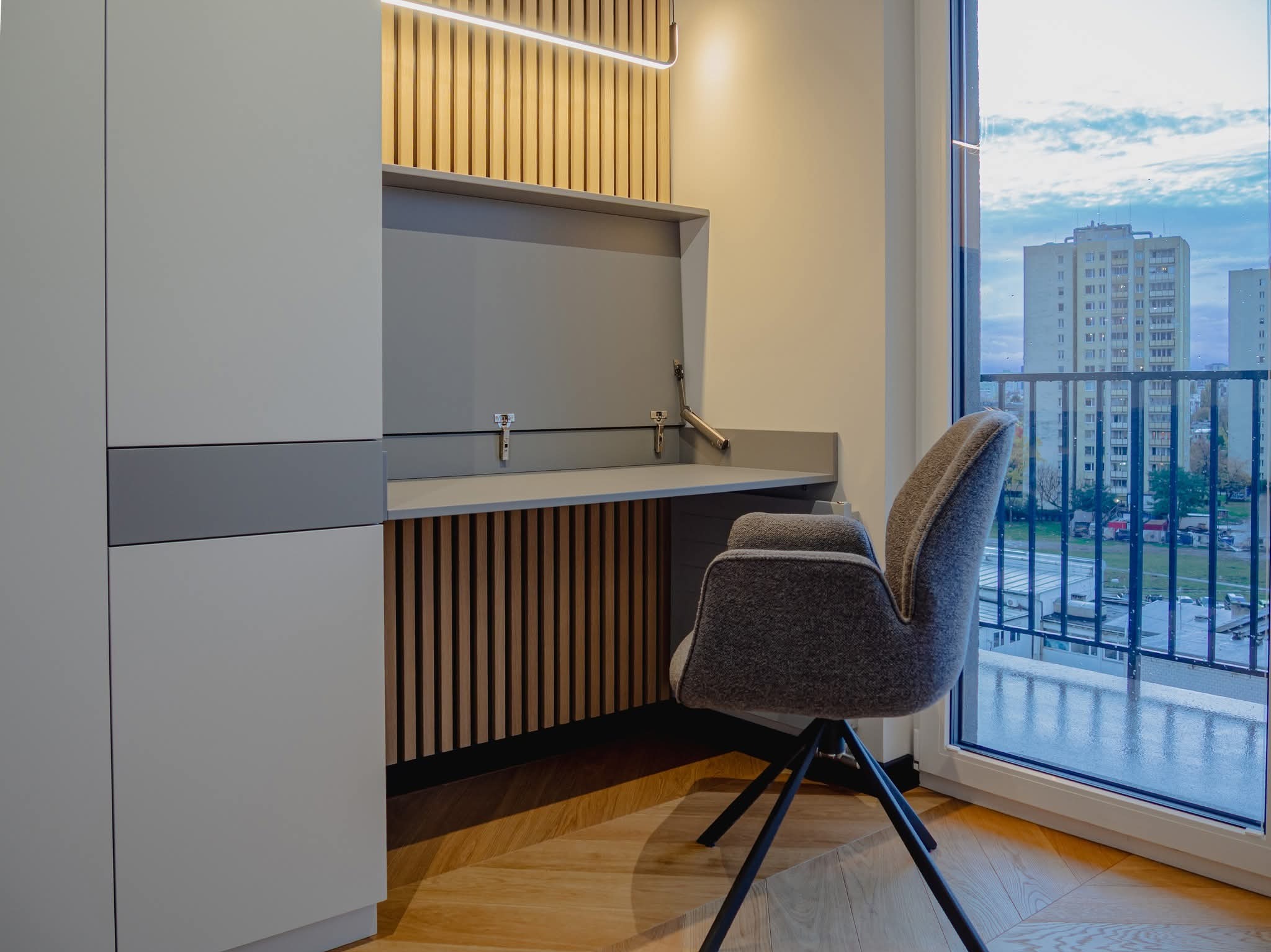 Nowoczesny pokój z biurkiem w zabudowie, szarym krzesłem i widokiem na miasto przez okno. Drewniane lamele i oświetlenie LED dodają wnętrzu ciepła.