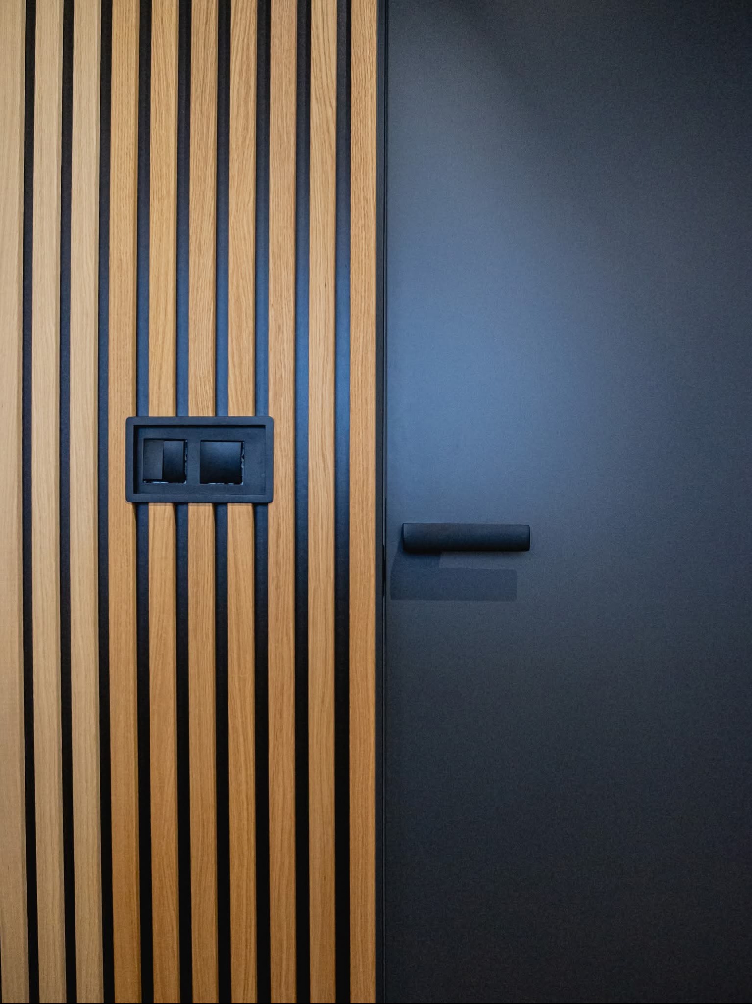 Panel ścienny z drewnianych listew w odcieniach brązu, z czarnymi wstawkami i gniazdkiem elektrycznym, sąsiadujący z gładkimi, grafitowymi drzwiami z minimalistyczną klamką.