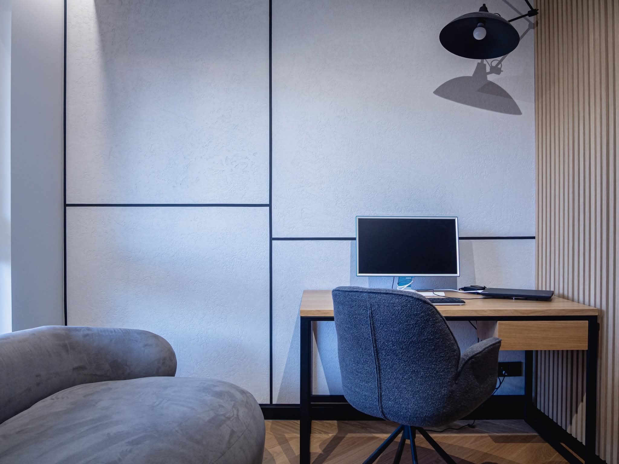 Nowoczesne wnętrze biurowe z teksturowaną ścianą, drewnianym biurkiem z czarną ramą, szarym fotelem i lampą. Minimalistyczny design z akcentami drewna.