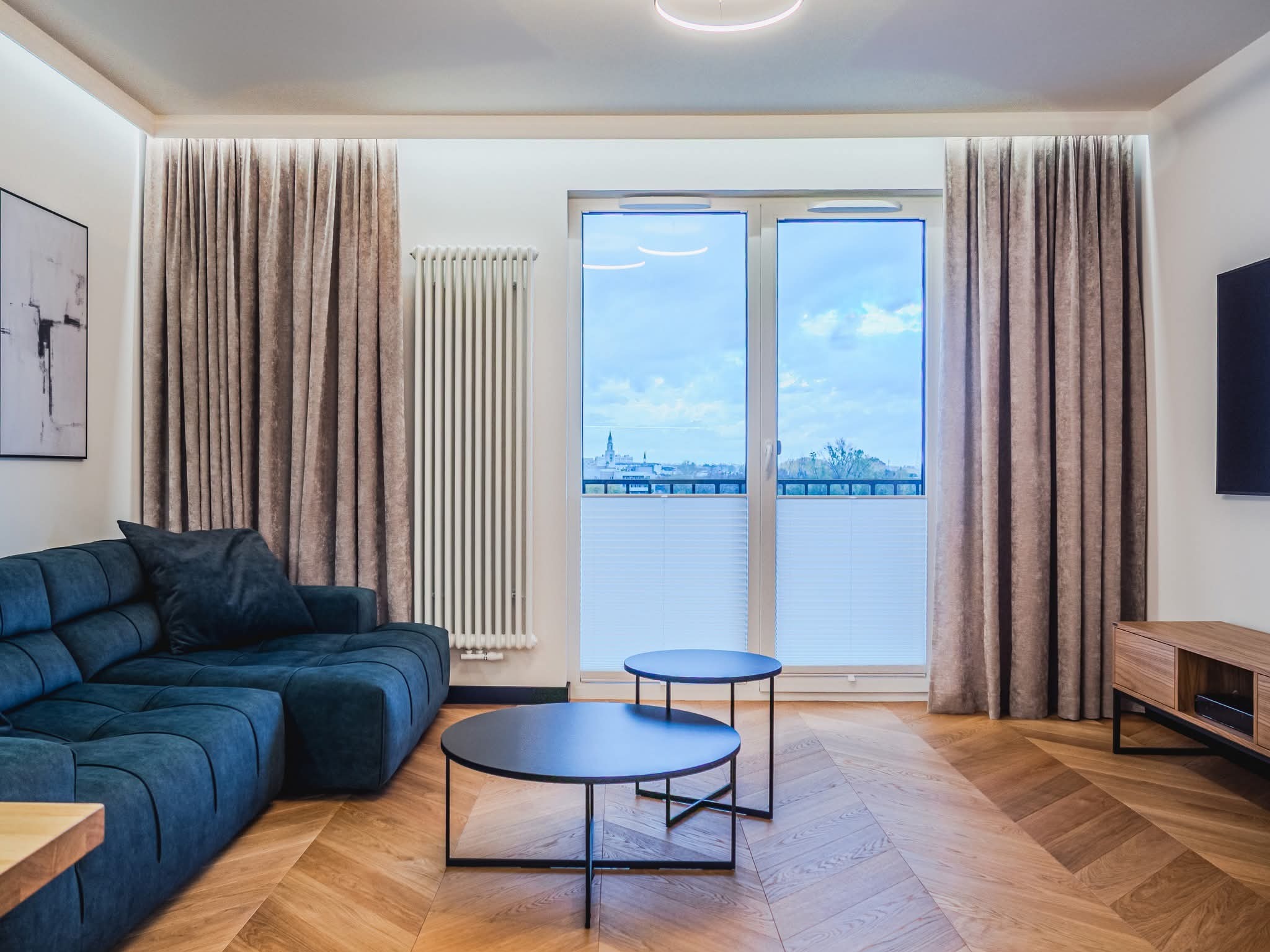 Salon z parkietem w jodełkę, niebieską sofą i okrągłymi stolikami. Widok na miasto przez okno z plisowanymi roletami. Nowoczesne oświetlenie i minimalistyczny wystrój.