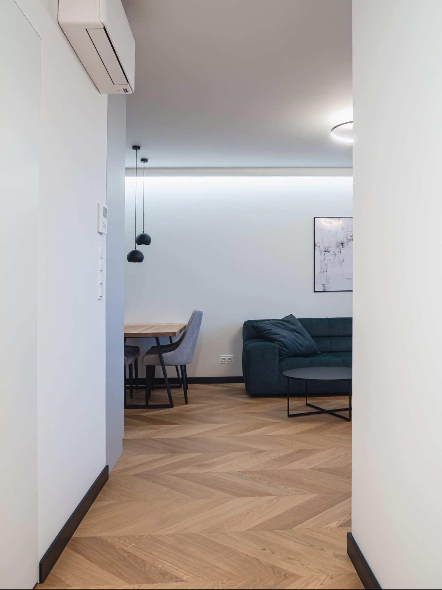Jasne parkietowe panele ułożone w jodełkę, widok z korytarza na salon z sofą i jadalnię z czarnymi lampami. Minimalistyczne wnętrze w odcieniach bieli i szarości.
