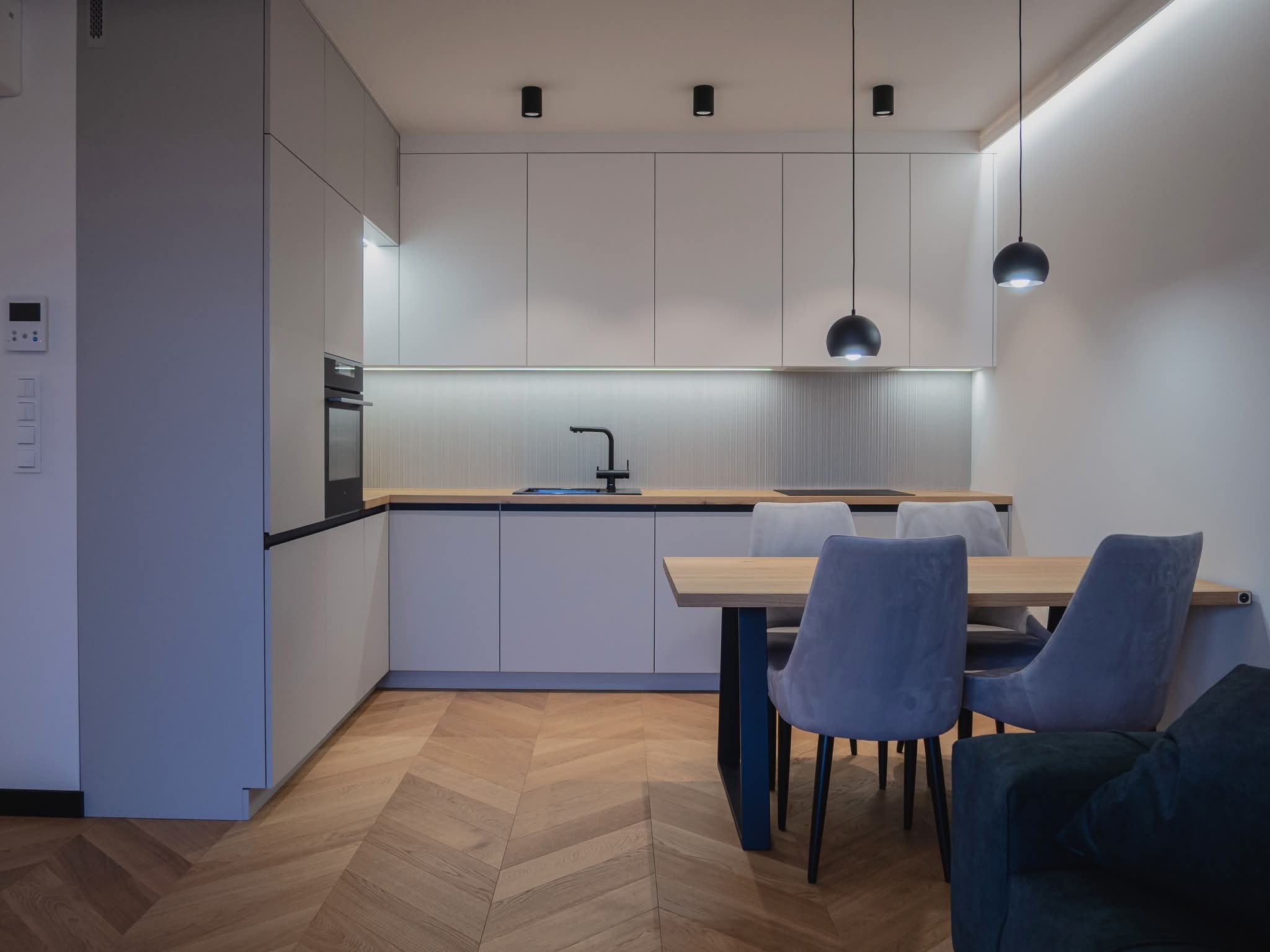 Nowoczesna kuchnia z szarymi szafkami, drewnianym blatem i parkietem w jodełkę. Czarne lampy wiszące nad stołem z krzesłami w odcieniach szarości.