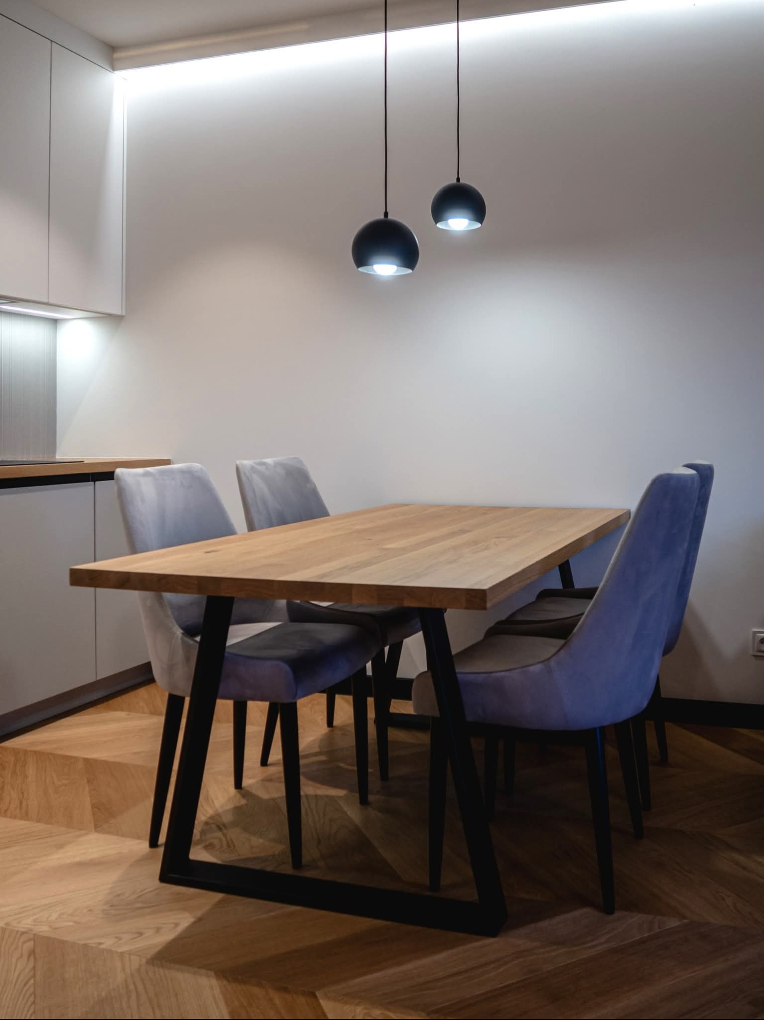 Nowoczesna kuchnia z drewnianym stołem i szarymi krzesłami na parkiecie ułożonym we wzór jodełki. Dwie czarne lampy wiszą nad stołem, a oświetlenie LED podkreśla minimalistyczny design.
