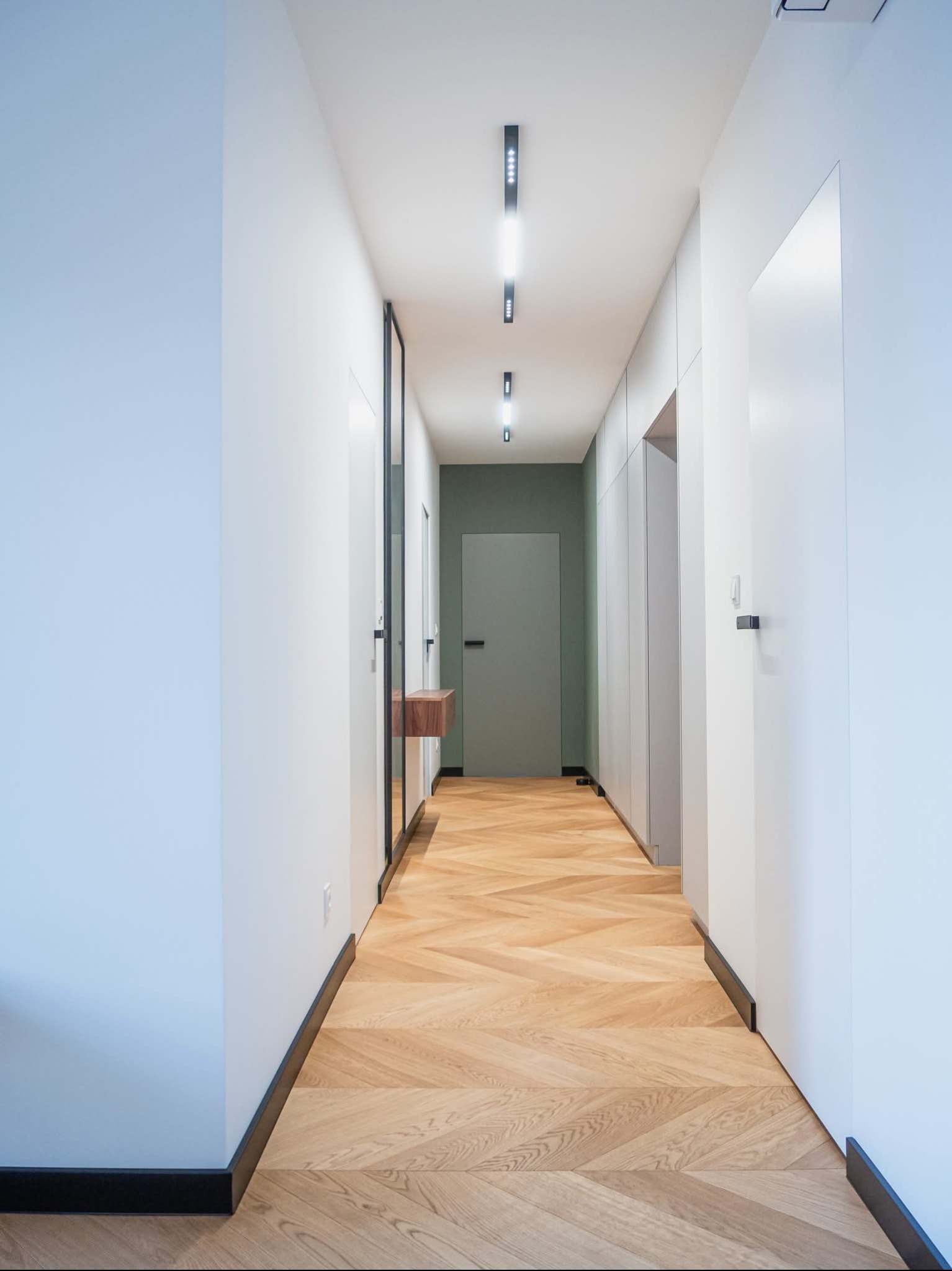 Nowoczesny korytarz z parkietem ułożonym w jodełkę, białe ściany i szafy w zabudowie, oświetlenie punktowe. Minimalistyczny design z akcentami drewna.