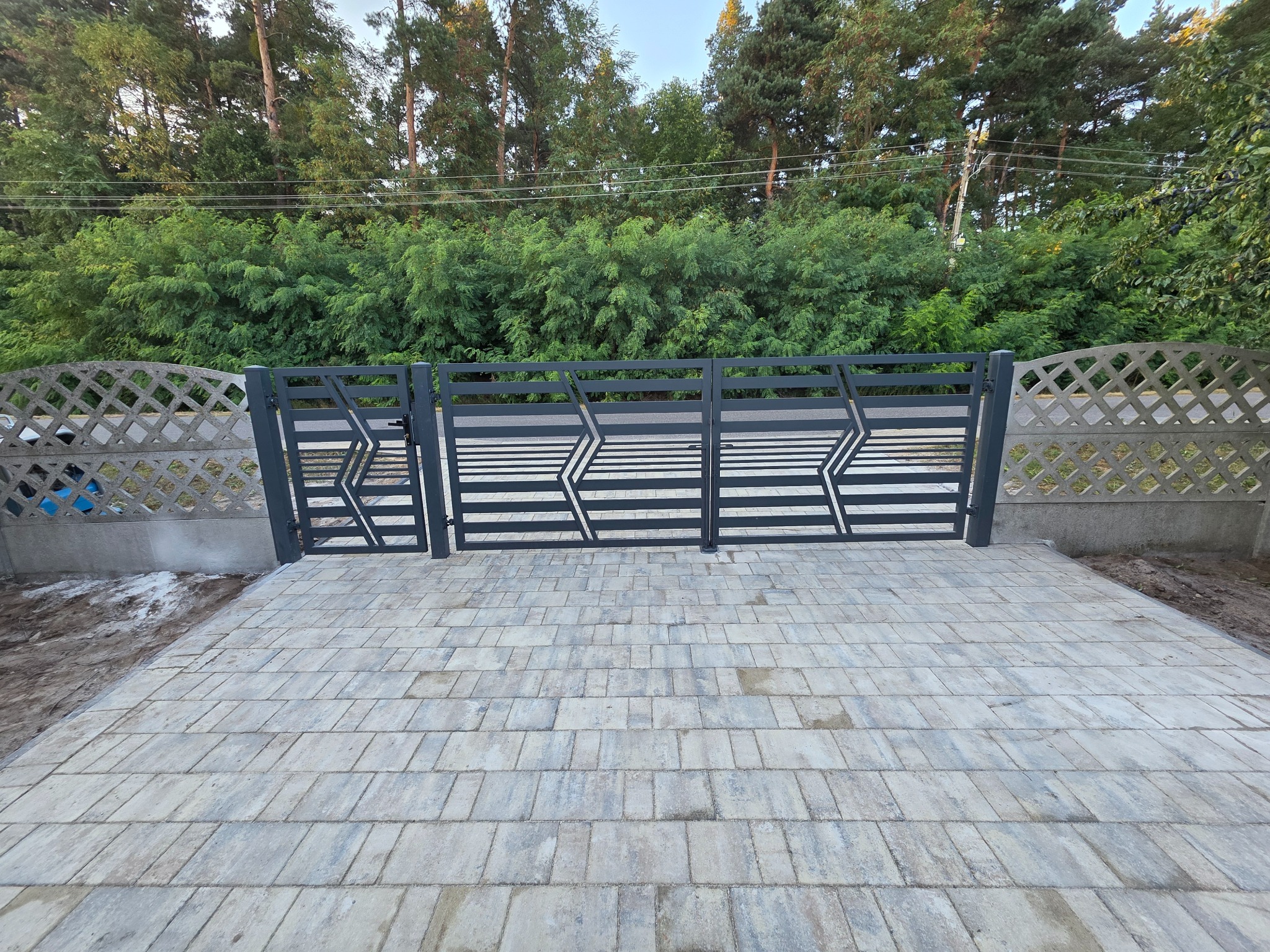Nowoczesna, grafitowa brama wjazdowa z geometrycznym wzorem, uzupełniona betonowym ogrodzeniem z ażurowym wypełnieniem. Kostka brukowa na podjeździe.