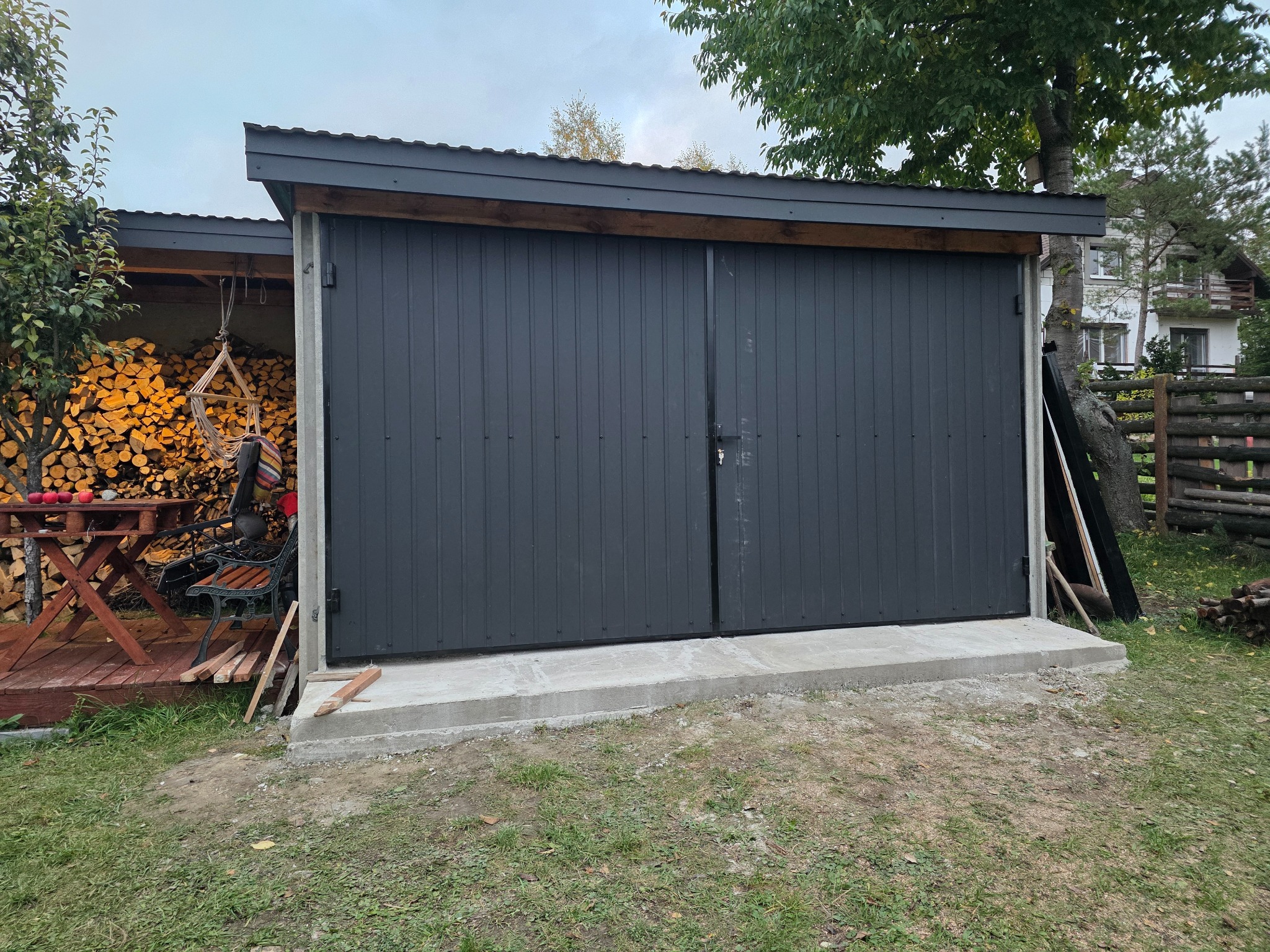 Nowoczesna, grafitowa brama garażowa z pionowymi panelami, osadzona na betonowej podstawie. W tle widoczny skład drewna i elementy drewnianej konstrukcji.