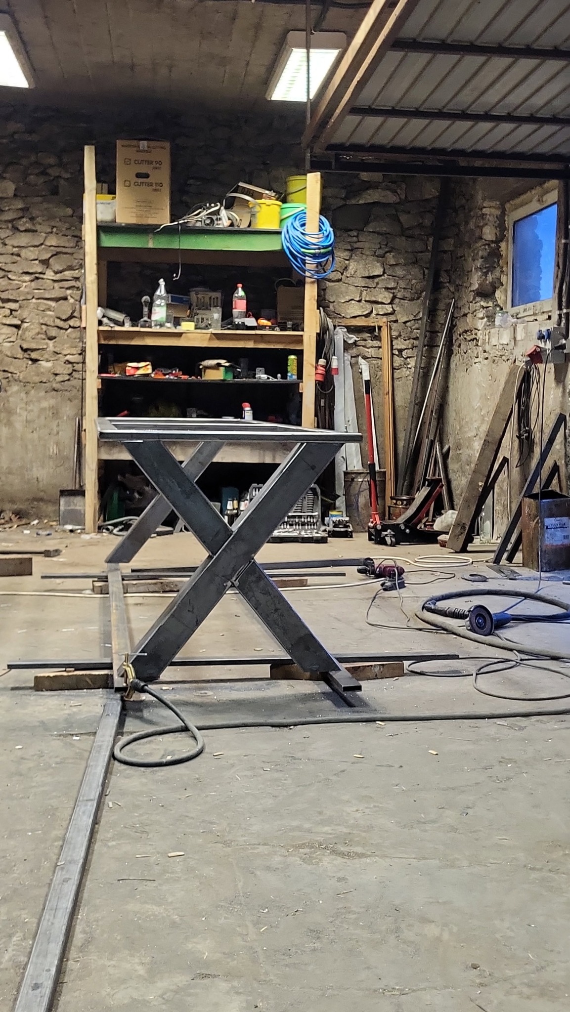 Metalowa konstrukcja stołu z nogami w kształcie litery X, spawana w warsztacie z kamiennymi ścianami i narzędziami w tle. Surowy, industrialny charakter.