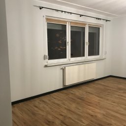 DAR-HAUS DARIUSZ NOWAK USŁUGI OGÓLNOBUDOWLANE - Jasne panele podłogowe w pokoju z białymi ścianami, grzejnikiem i oknem z roletami. Czarne listwy przypodłogowe. Minimalistyczne wnętrze po remoncie.