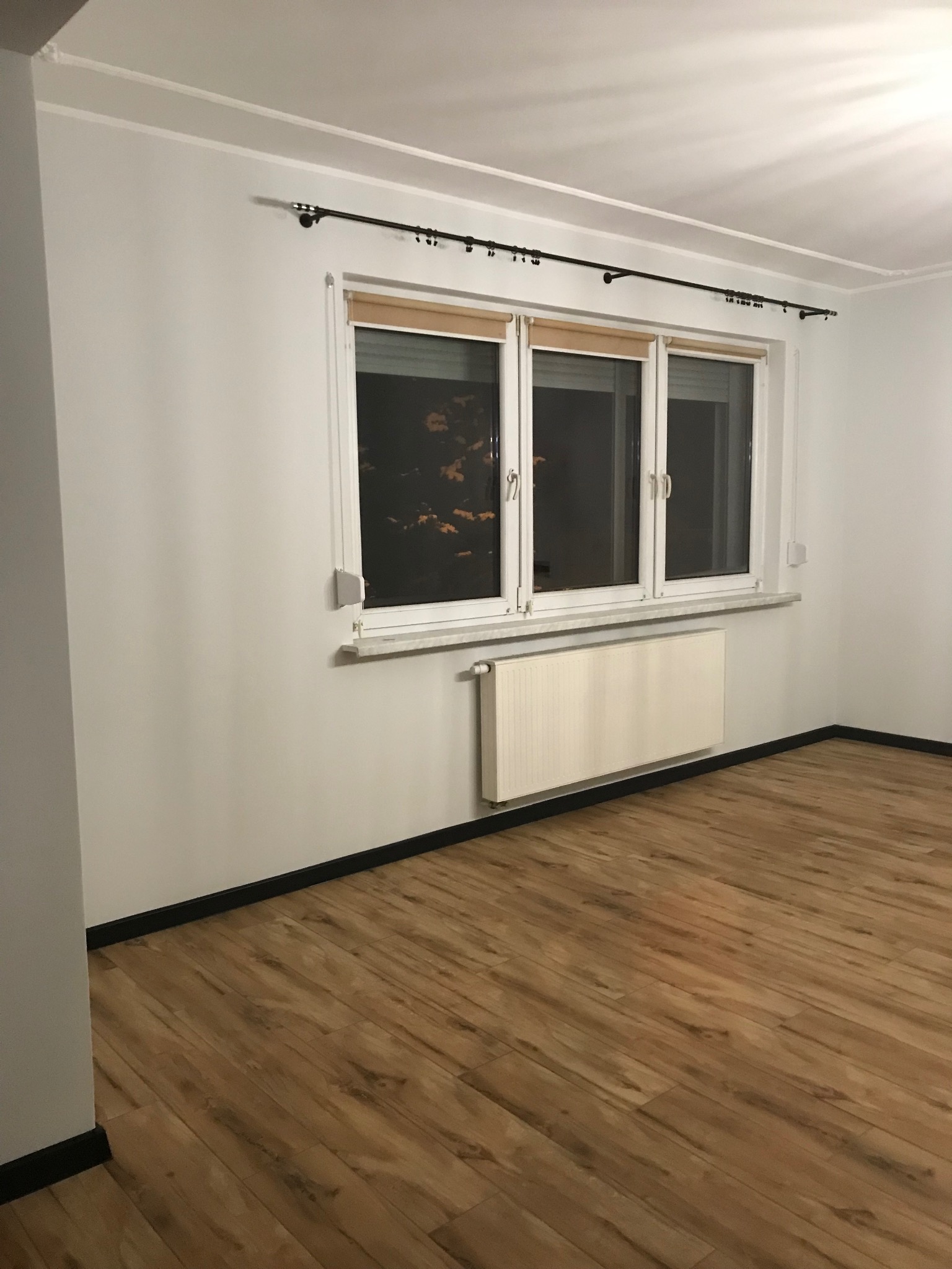 Jasne panele podłogowe w pokoju z białymi ścianami, grzejnikiem i oknem z roletami. Czarne listwy przypodłogowe. Minimalistyczne wnętrze po remoncie.