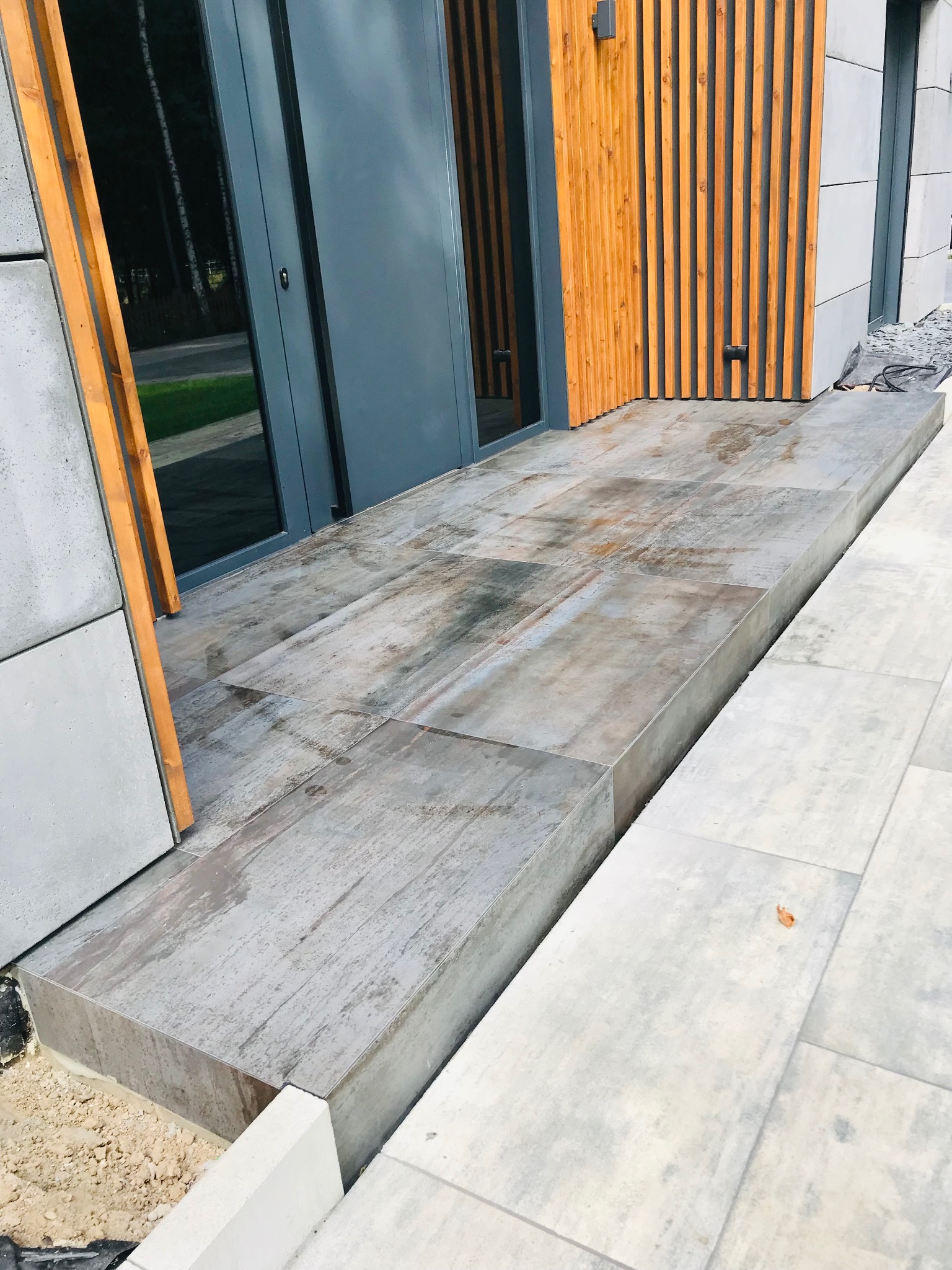 Nowoczesne wykończenie wejścia do domu z szarymi płytkami imitującymi drewno, drewnianą elewacją i grafitowymi drzwiami. Widoczne detale wykończeniowe.