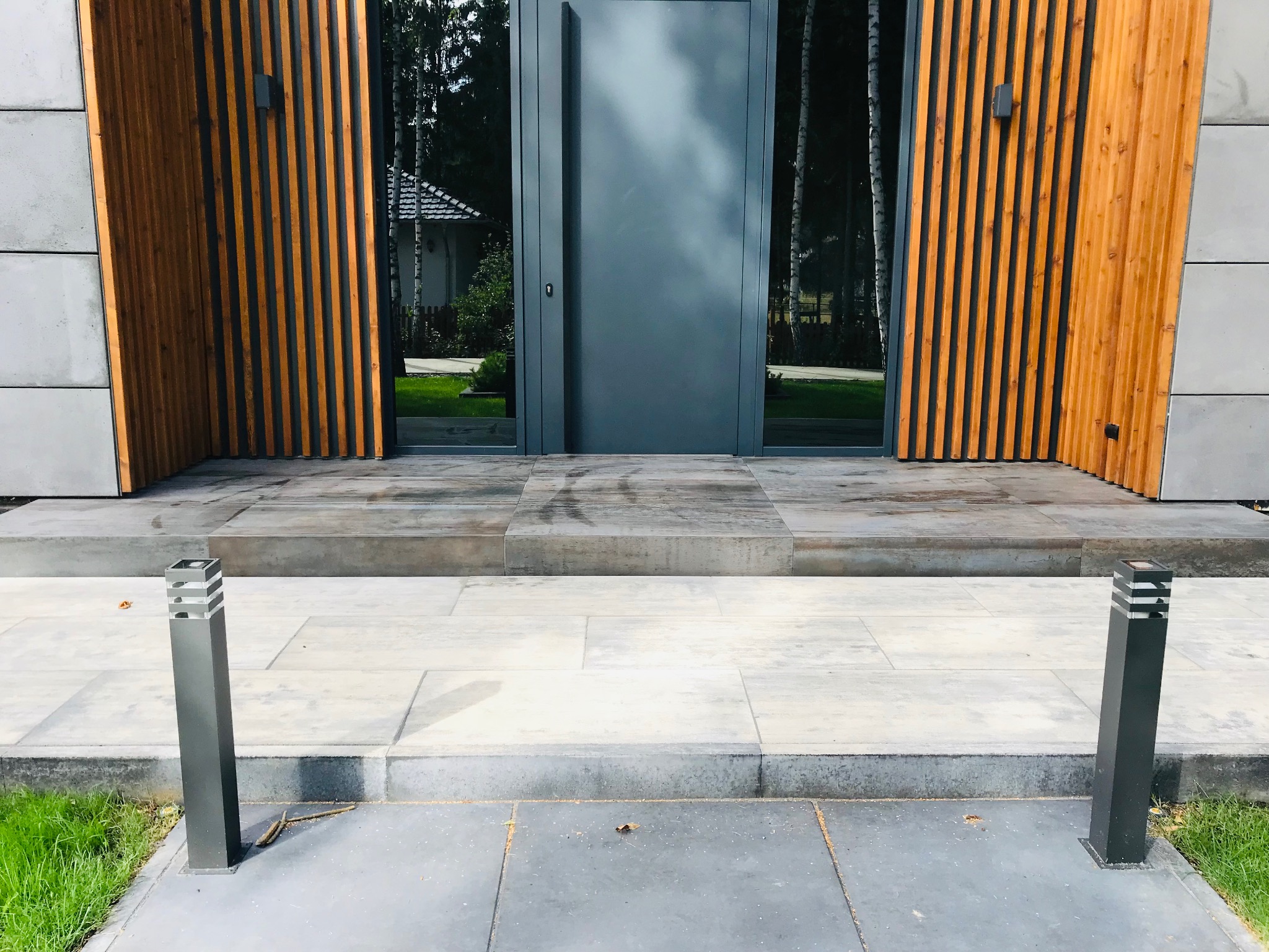 Nowoczesne wejście do domu z szarą, minimalistyczną stolarką drzwiową, drewnianymi panelami i kamienną posadzką. Oświetlenie zewnętrzne w postaci słupków.