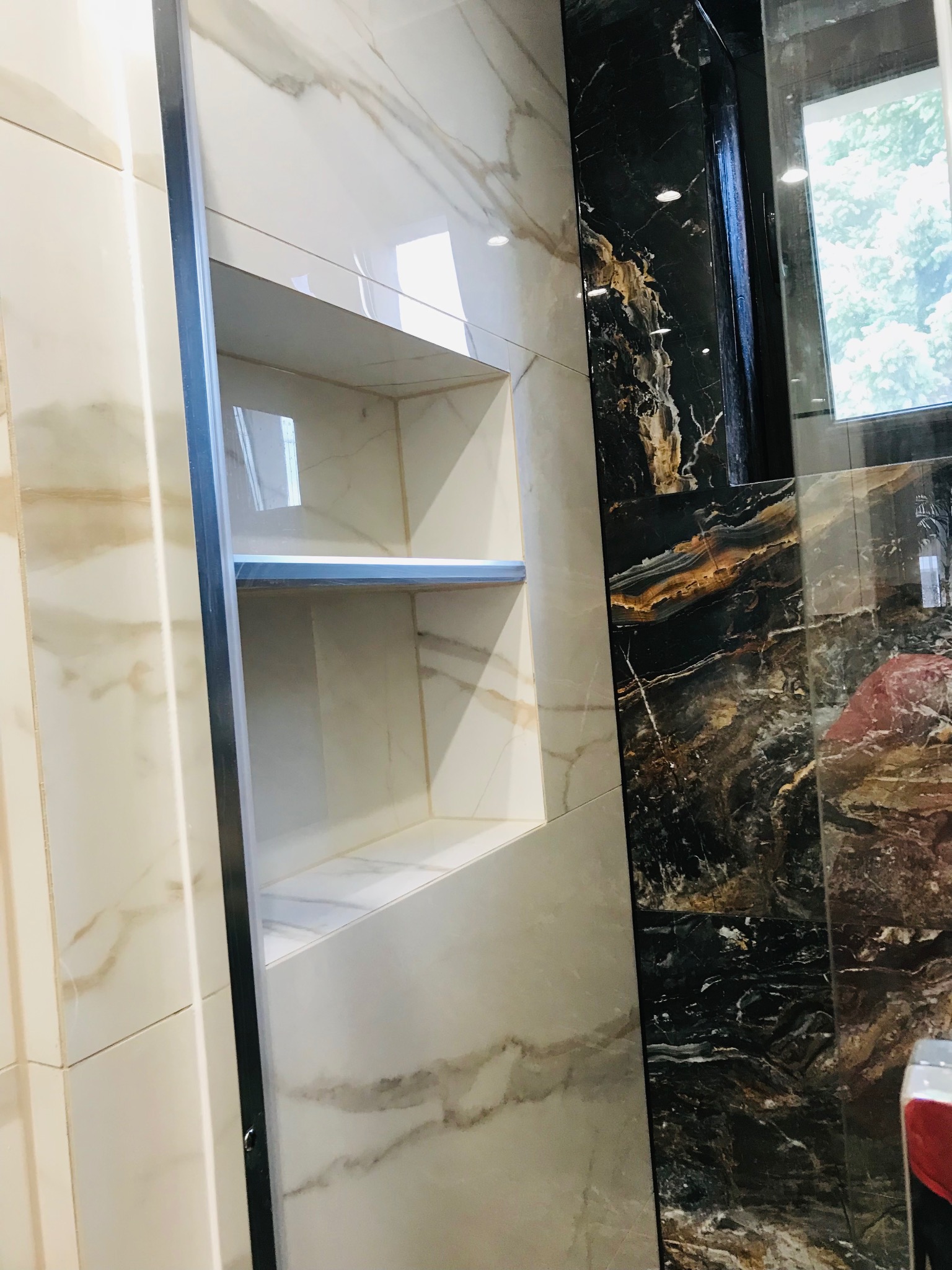 Wnęka w ścianie wyłożona płytkami imitującymi marmur, z półkami. Obok ściana z czarnych płytek z wyraźnym, złotym żyłowaniem. Nowoczesna aranżacja łazienki.