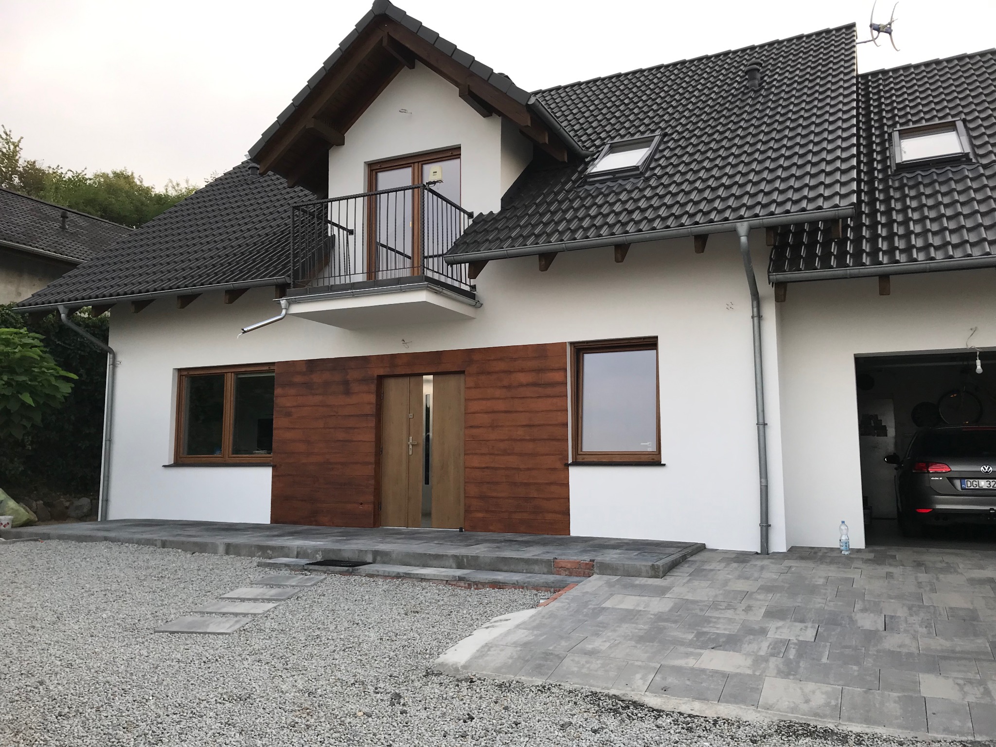 Nowoczesny dom jednorodzinny z białymi ścianami, drewnianą elewacją i ciemną dachówką. Widoczny balkon, okna z drewnianą stolarką i szara kostka brukowa.