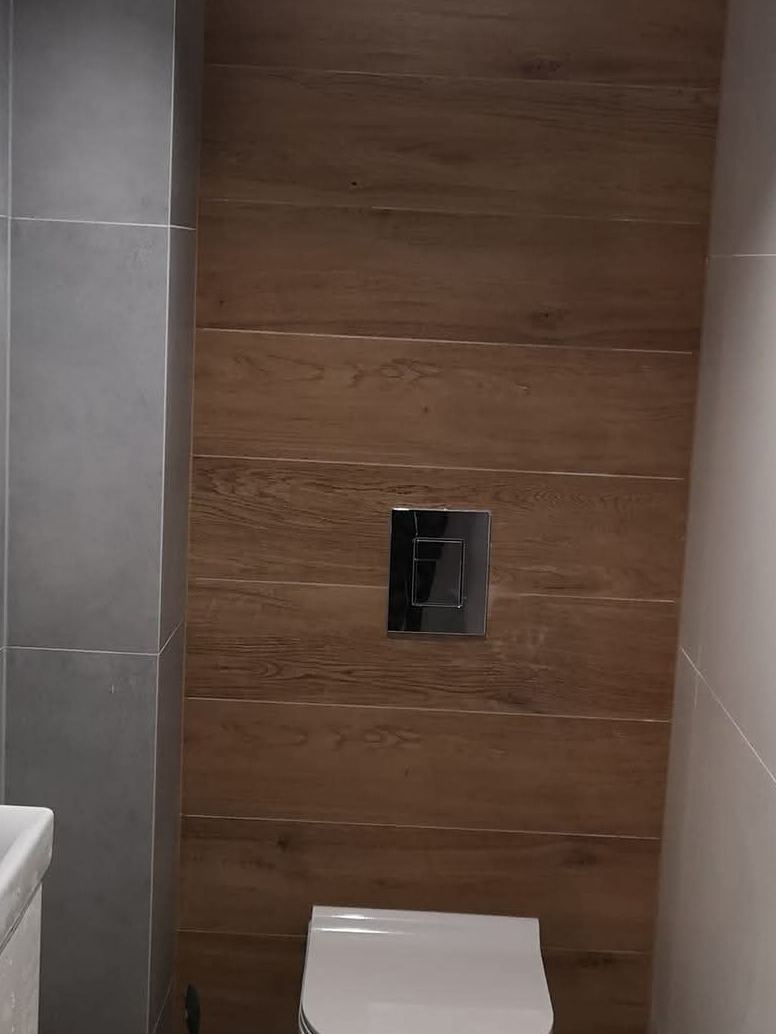 Nowoczesna toaleta z drewnopodobnymi płytkami na ścianie, chromowany przycisk spłuczki i szare płytki po lewej stronie. Minimalistyczny design.