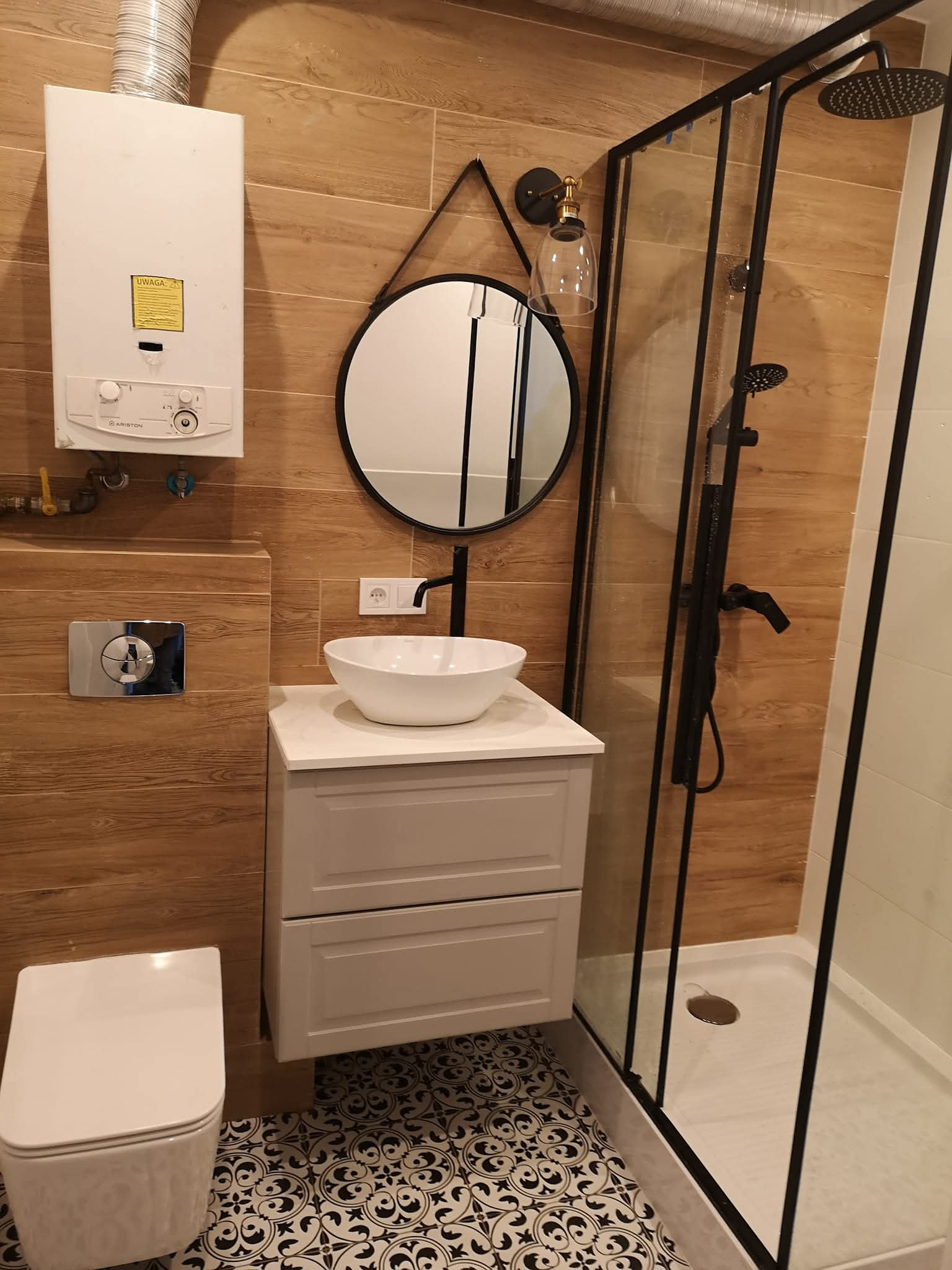 Nowoczesna łazienka z kabiną prysznicową typu walk-in, umywalką nablatową i okrągłym lustrem. Płytki imitujące drewno na ścianach, wzorzysta podłoga.