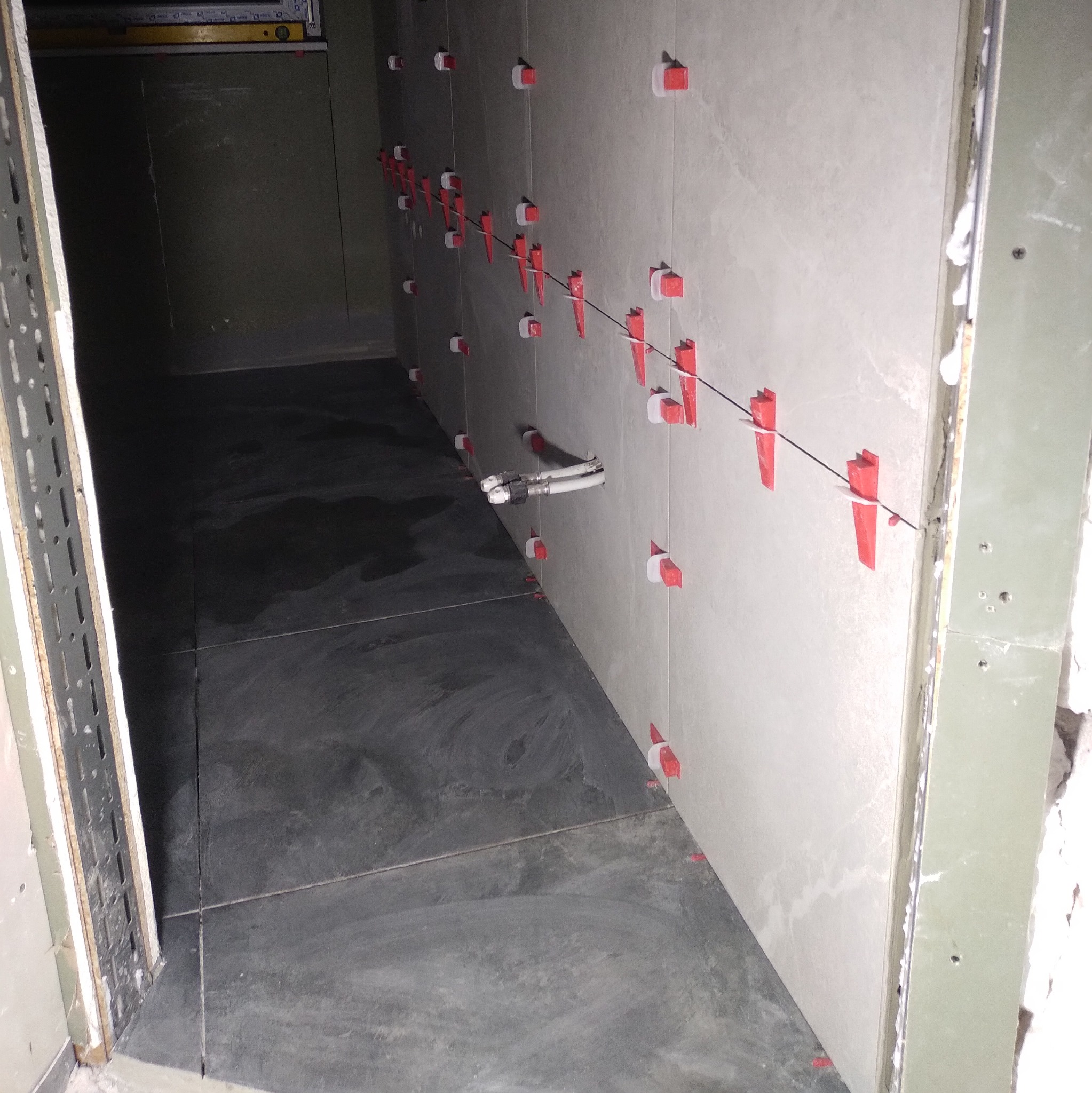 Ściana i podłoga w trakcie układania płytek, z użyciem systemu poziomowania. Widoczne białe płytki na ścianie i ciemne na podłodze, czerwone kliny i elementy dystansowe.