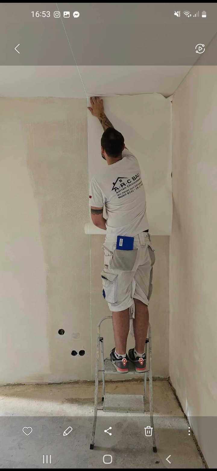 Pracownik na drabinie montuje tapetę na ścianie w pomieszczeniu w trakcie remontu. Widoczny laser wyznaczający pion. Na ścianie ślady po instalacji elektrycznej.