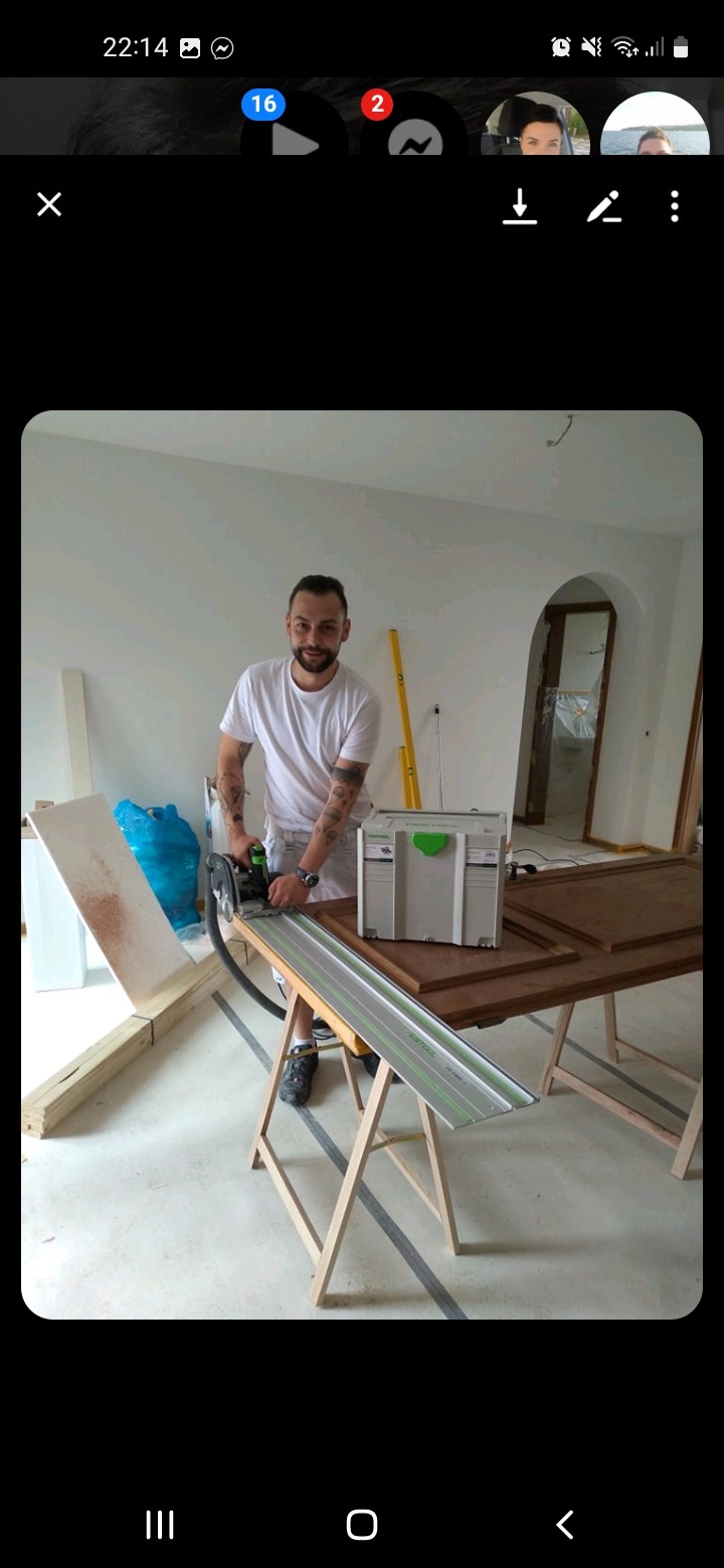 Uśmiechnięty mężczyzna z tatuażami tnie deskę piłą na stole warsztatowym w jasnym wnętrzu. Widoczny poziomica i łukowate przejście do innego pomieszczenia.