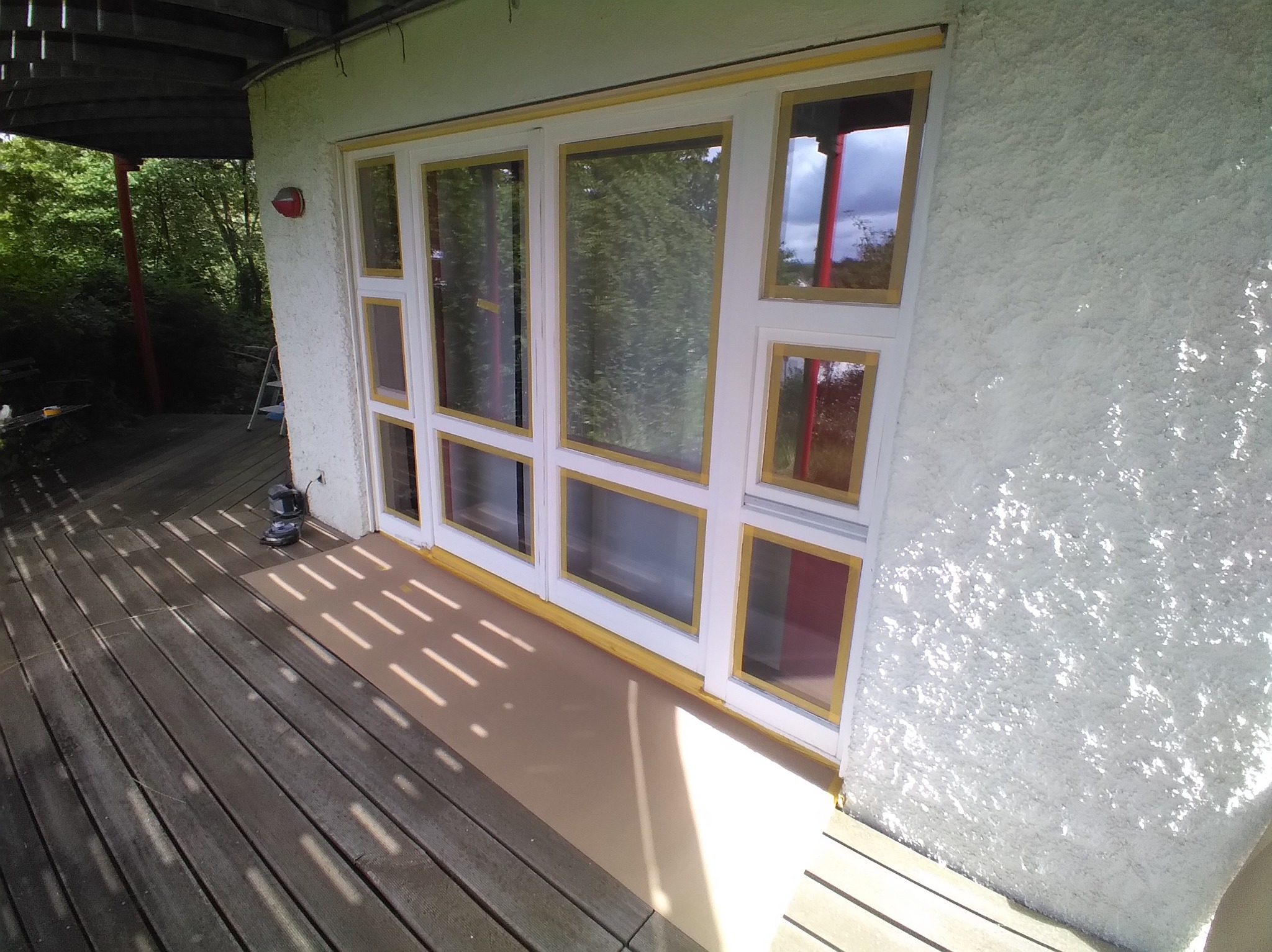 Biała, teksturowana ściana z oknami w białych ramach, zabezpieczonymi żółtą taśmą malarską. Widoczny fragment drewnianego tarasu i zieleń w tle.