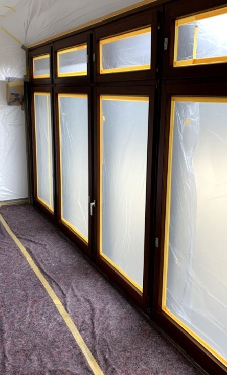 Przygotowane do malowania okna z brązowymi ramami, zabezpieczone folią i taśmą malarską, podłoga zabezpieczona folią ochronną. Widok z boku.