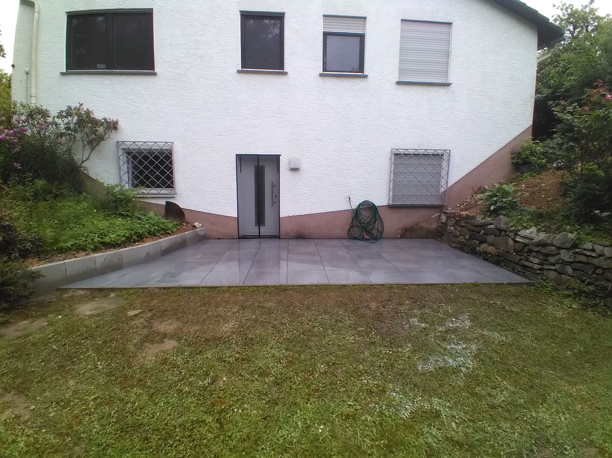 Nowoczesny taras z szarych płyt, przylegający do białego domu z ciemnymi oknami i zielonym ogrodem. Wąż ogrodowy zwinięty przy ścianie.