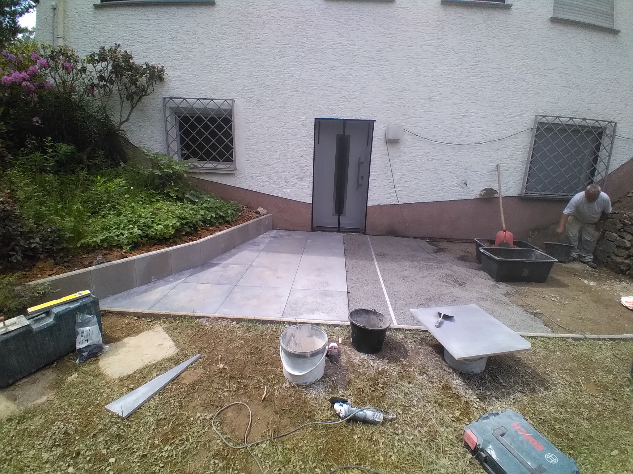 Układanie szarych płyt chodnikowych na zewnątrz budynku. Widoczne narzędzia, wiadro, poziomica i pracownik w tle. Częściowo ukończona praca.