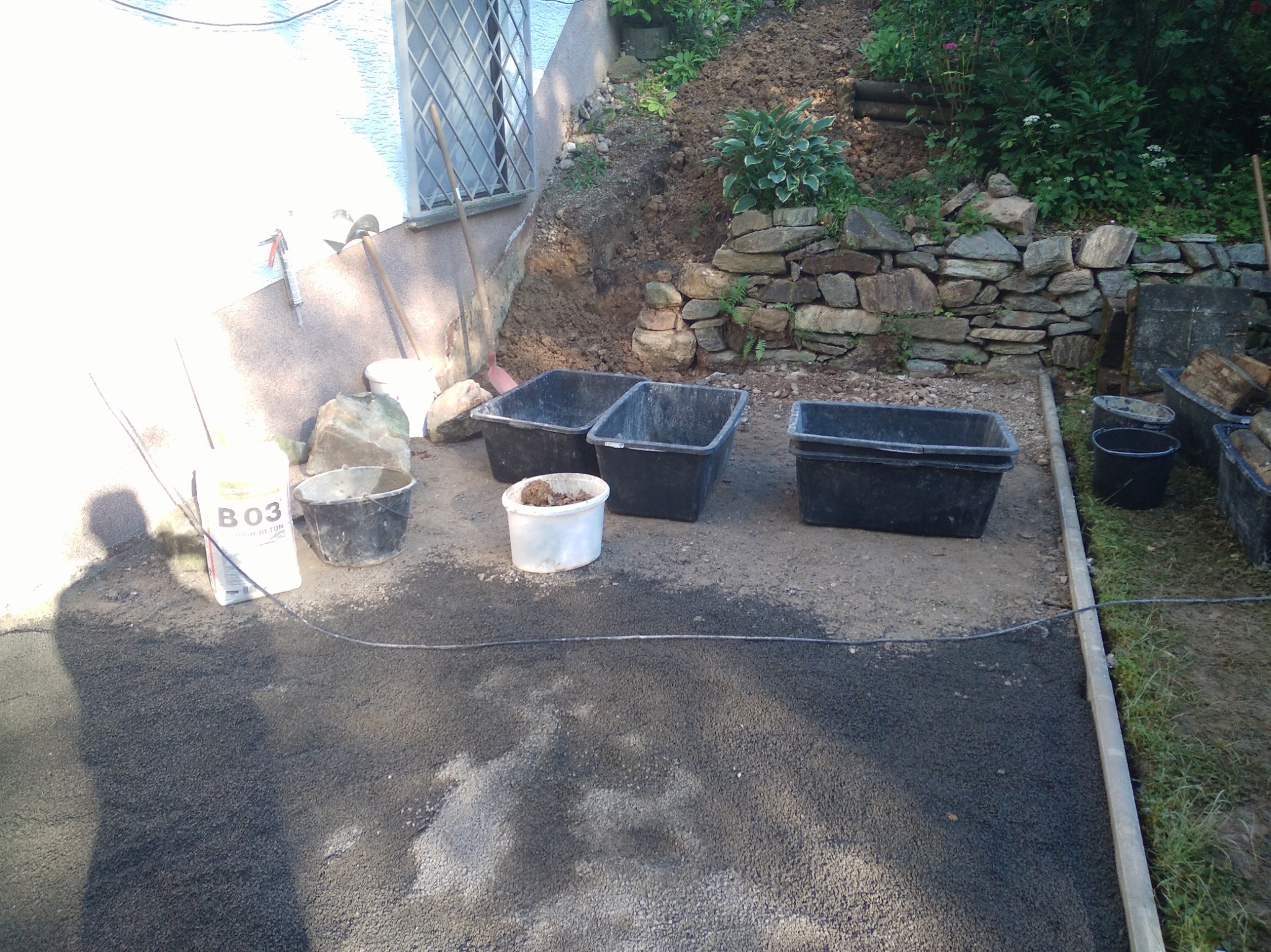 Przygotowanie terenu: cztery czarne pojemniki, wiadro, worek z betonem B03, narzędzia opierają się o ścianę. W tle ziemia i kamienny murek.