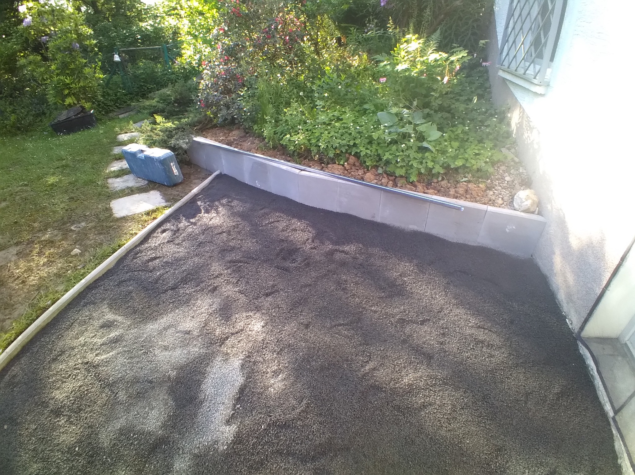 Przygotowanie podłoża z czarnego kruszywa pod taras lub chodnik, obrzeża z szarej kostki, widok z góry, fragment ogrodu w tle.