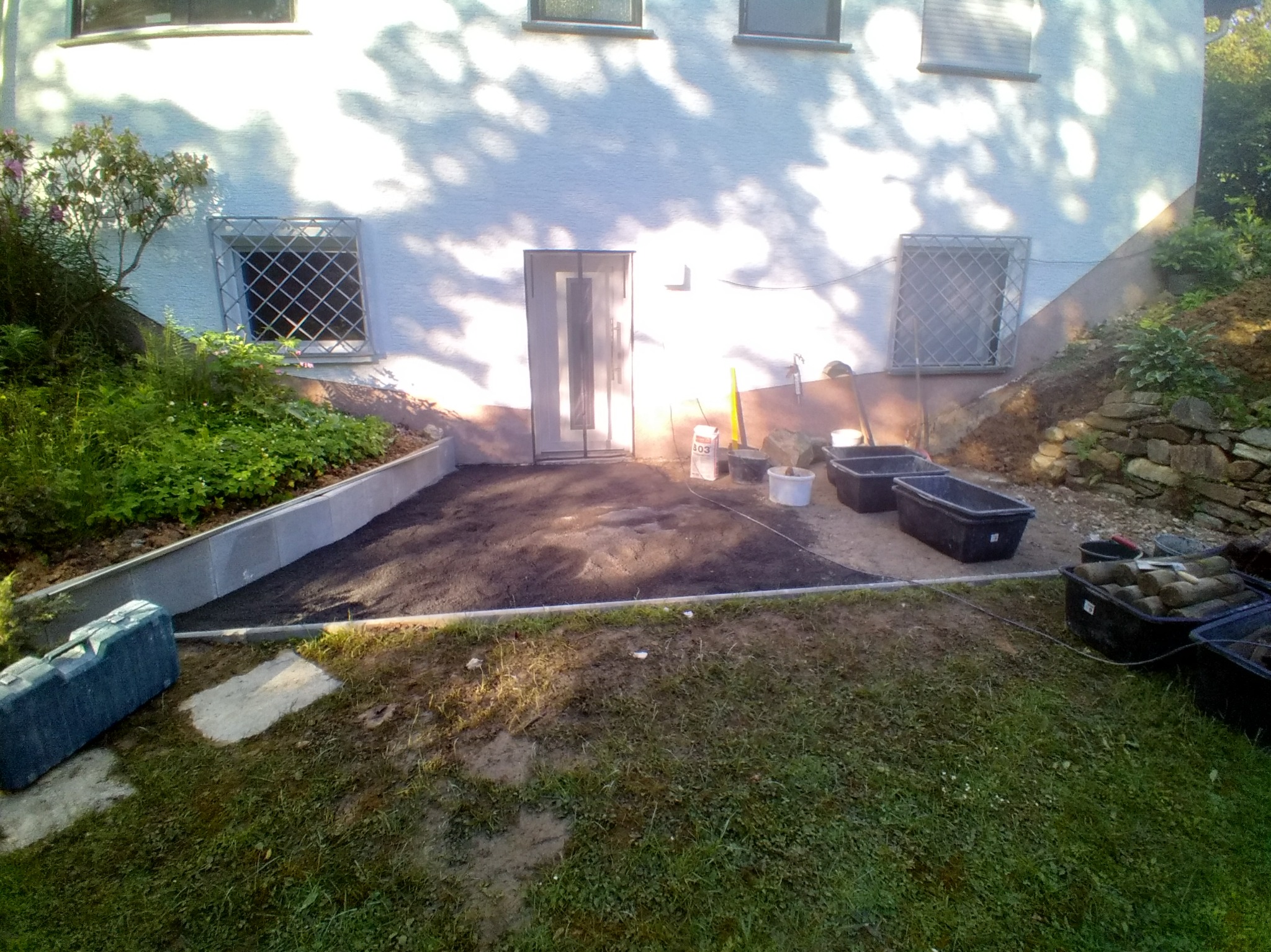 Przygotowanie podłoża pod taras w Olsztynie: wyrównany teren z widocznymi narzędziami i materiałami budowlanymi. Prace ziemne w toku, widok na ścianę budynku.