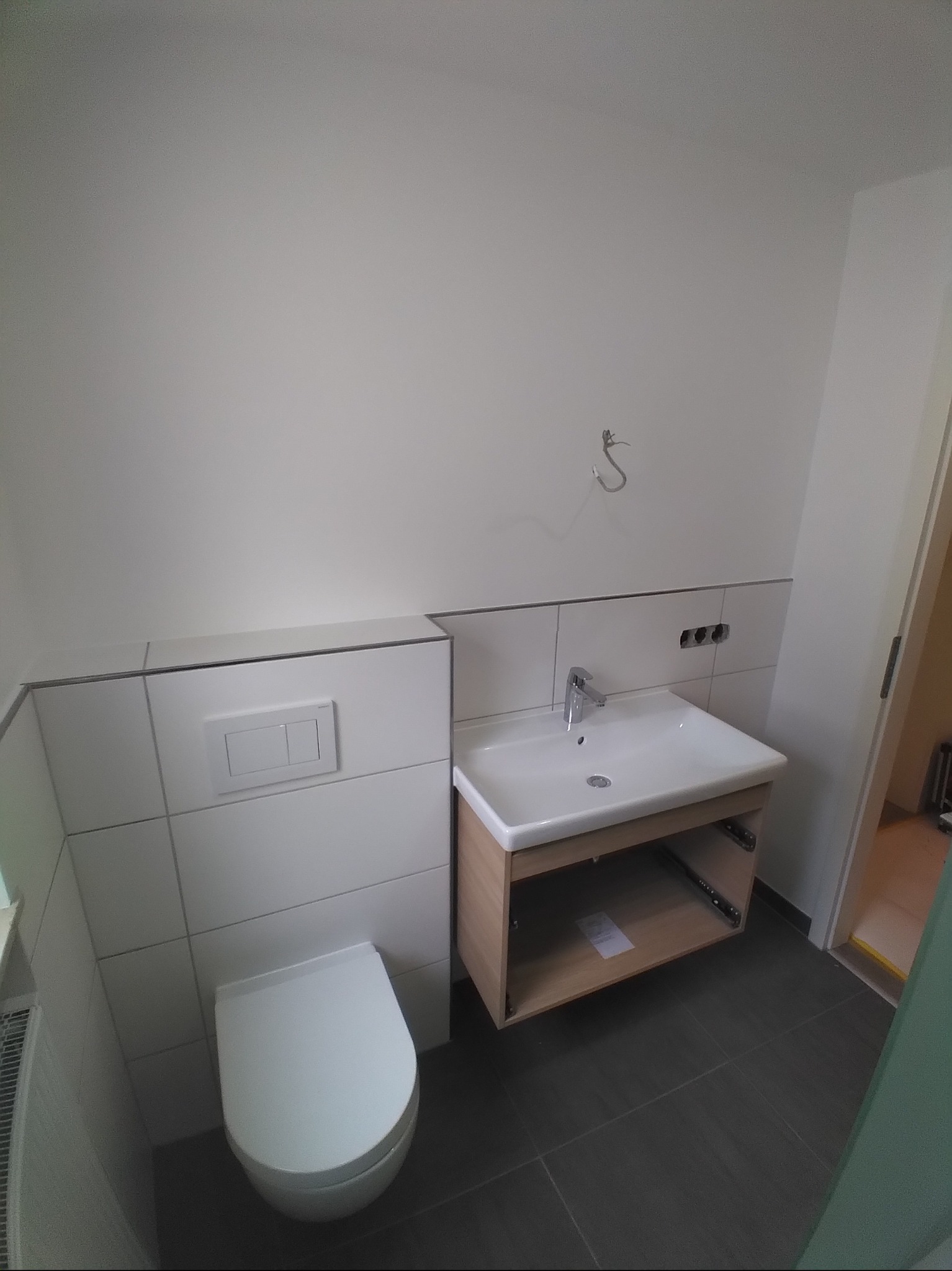 Nowoczesna łazienka z podwieszaną toaletą i umywalką na szafce, wykończona białymi płytkami i szarą podłogą. Widoczne elementy instalacji.