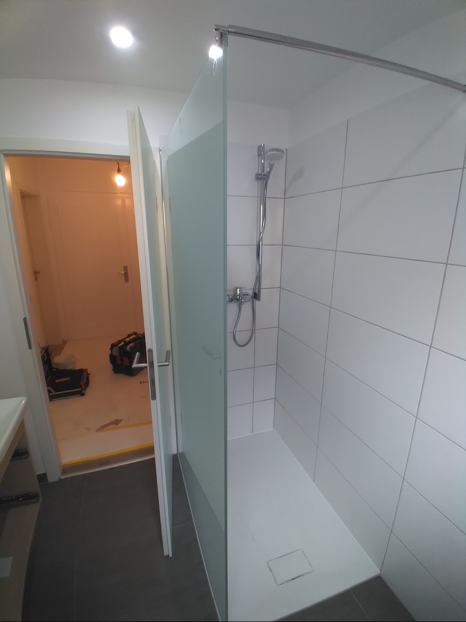 Nowoczesna kabina prysznicowa z białymi płytkami i szklaną ścianką, widok na otwarte drzwi i narzędzia w tle. Minimalistyczny design, szare płytki podłogowe.