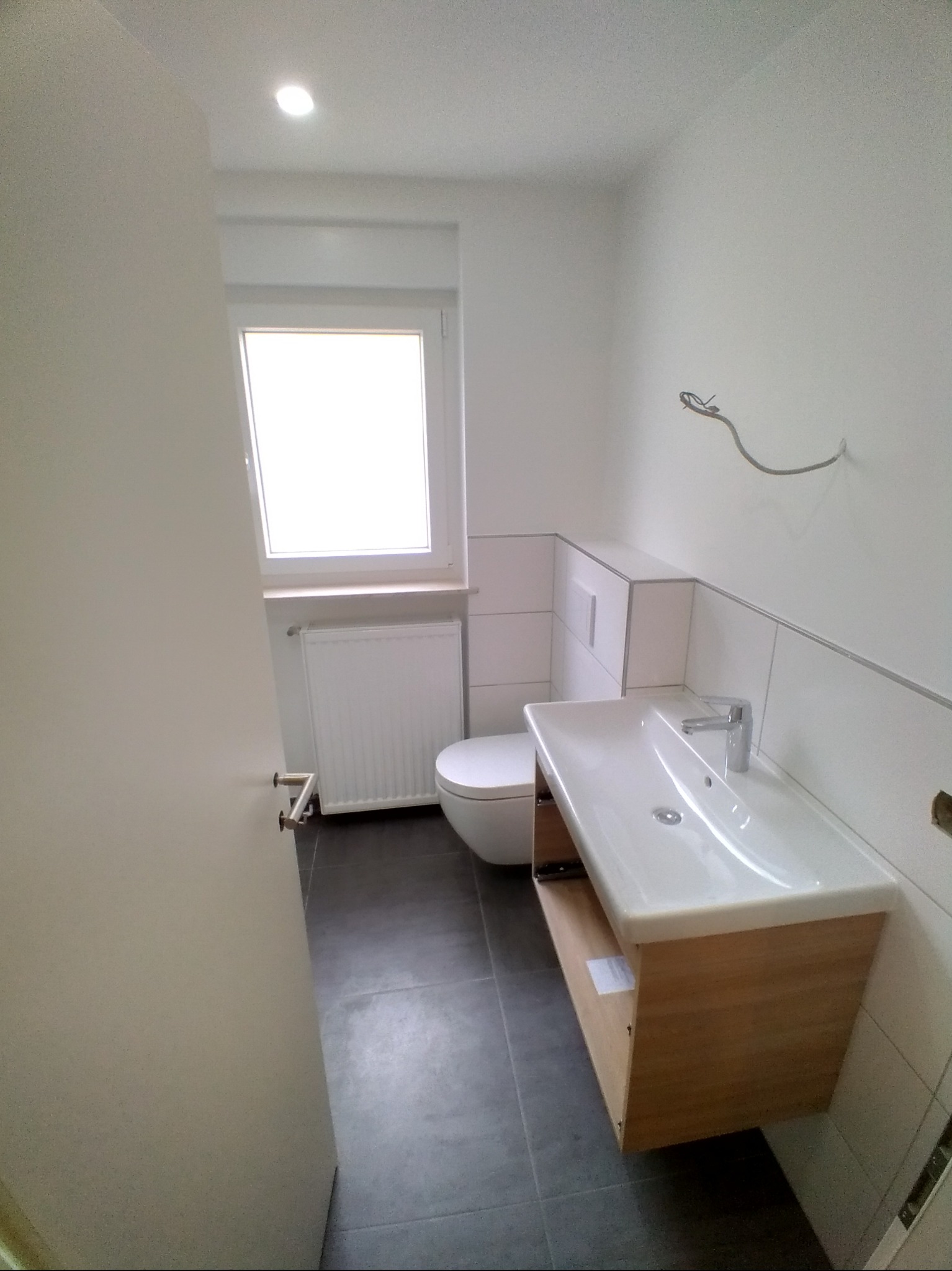 Jasna łazienka z umywalką na szafce z jasnego drewna, toaletą i grzejnikiem pod oknem. Widoczne białe płytki i szare płytki podłogowe. Minimalistyczny design.